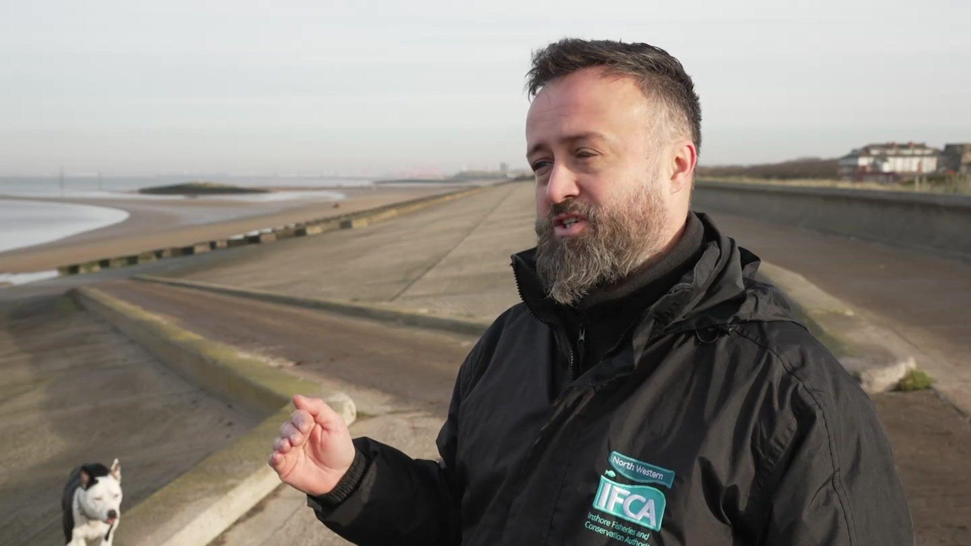 Mark Taylor from the North Western Inshore Fisheries and Conservation Authority stands near the breakwaters at Leasowe. A black and white dog stands in the background. 