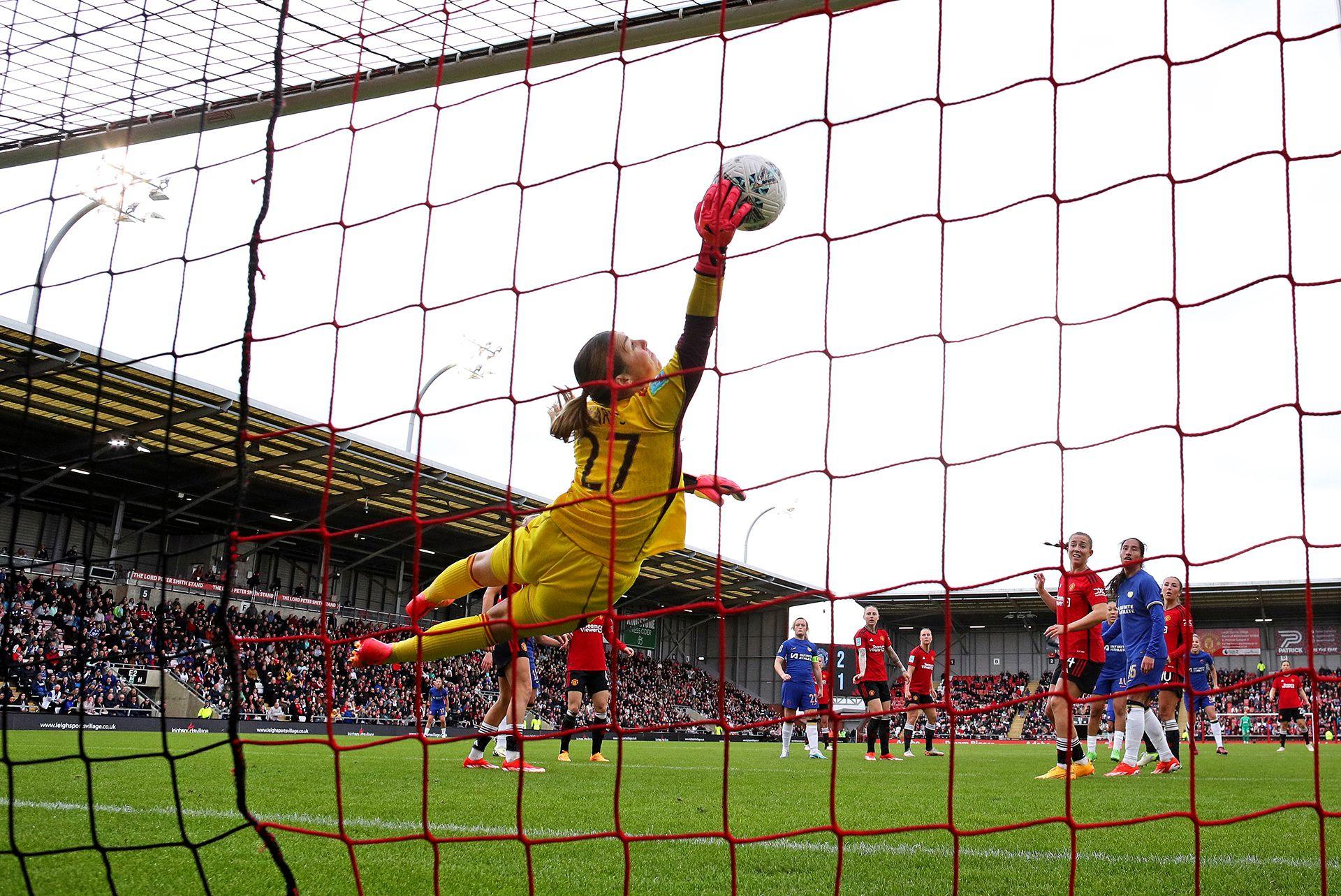 Mary Earps dives and saves the ball after Lauren James of Chelsea heads it towards the goal, she's diving in mid-air with a glove on the ball, while the photo is taken from the back of the goal showing the net in the foreground, and both Chelsea and Man Utd players in the background against the backdrop of the stadium and a grey sky, at Leigh Sports Village in April 2024.