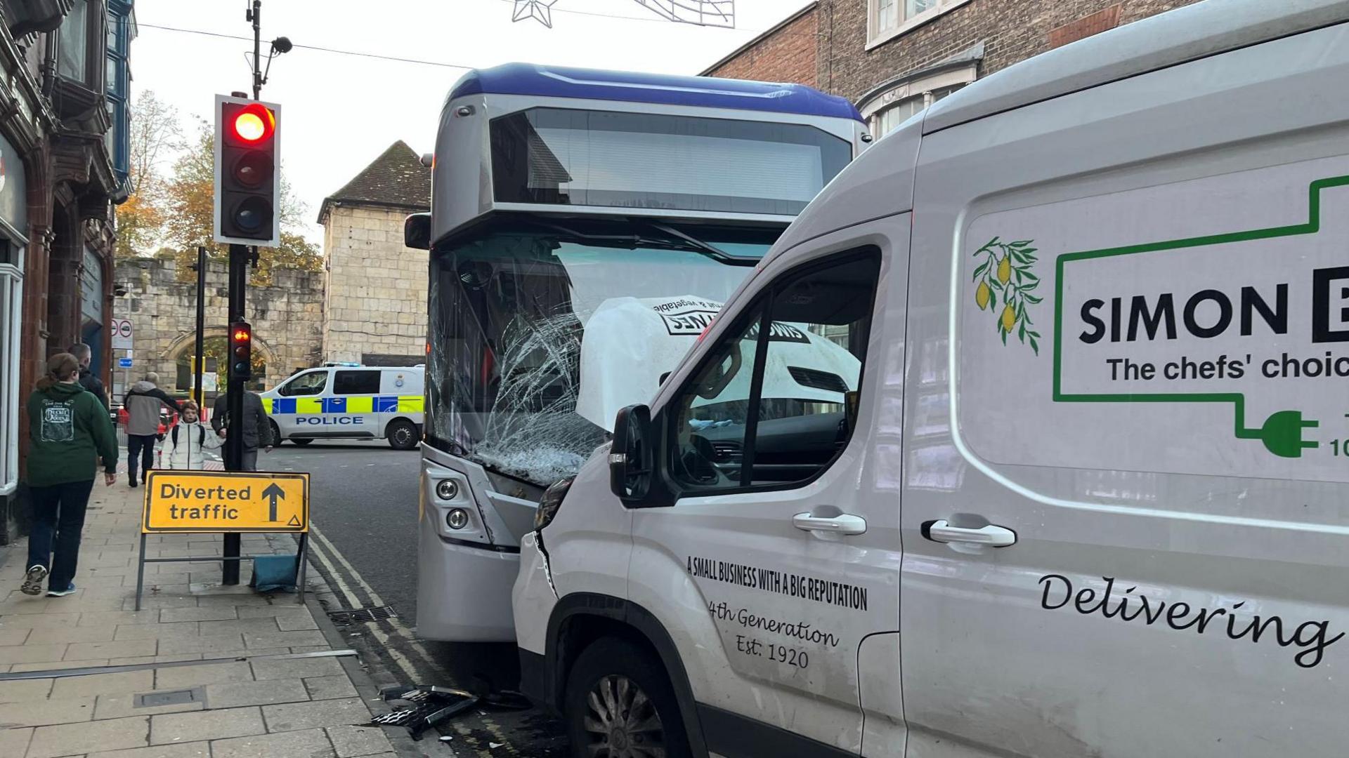 A white single decker bus crashed into the front of a white van. The bus has a cracked windscreen and the van has damage to its bonnet.