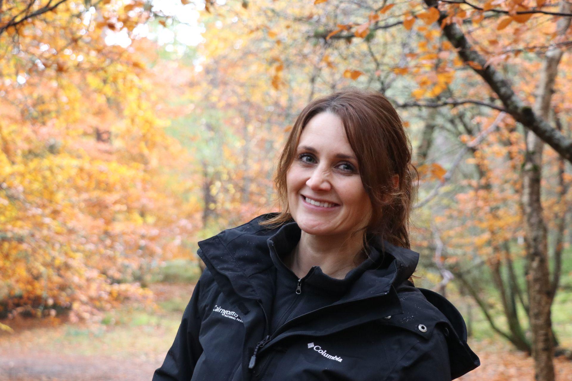 A woman, Sarah Henshall, smiles and looks into the camera. She has brown hair, pulled into a ponytail, and is wearing a heavy, black outdoor raincoat. She stands in front of a backdrop of trees in autumn. The leaves are a beautiful shade of gold and orange.