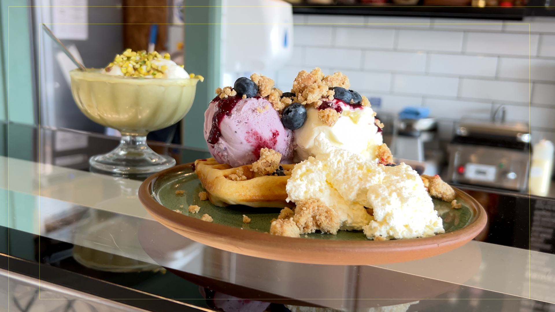 Two ice cream sundaes sit on a counter-top. One has vanilla and berry ice cream, with blueberries and crunchy topping. The other is green pistachio with expresso coffee.