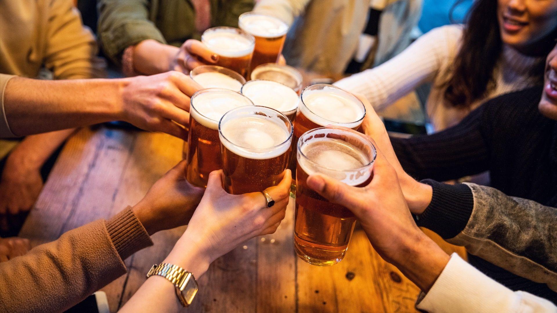 A group of people are toasting with pints of beer or lager around a wooden table. The photo is taken from above, focusing on the raised glasses and hands.