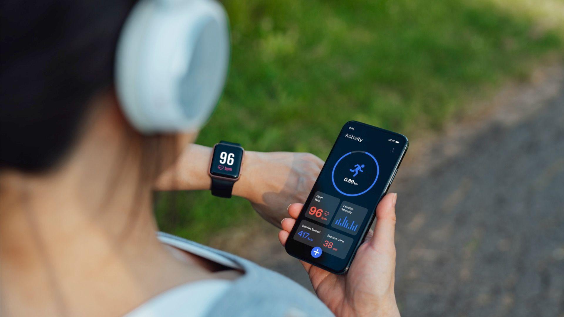 A person is holding a smartphone displaying a fitness tracking app while wearing white headphones and a smartwatch.