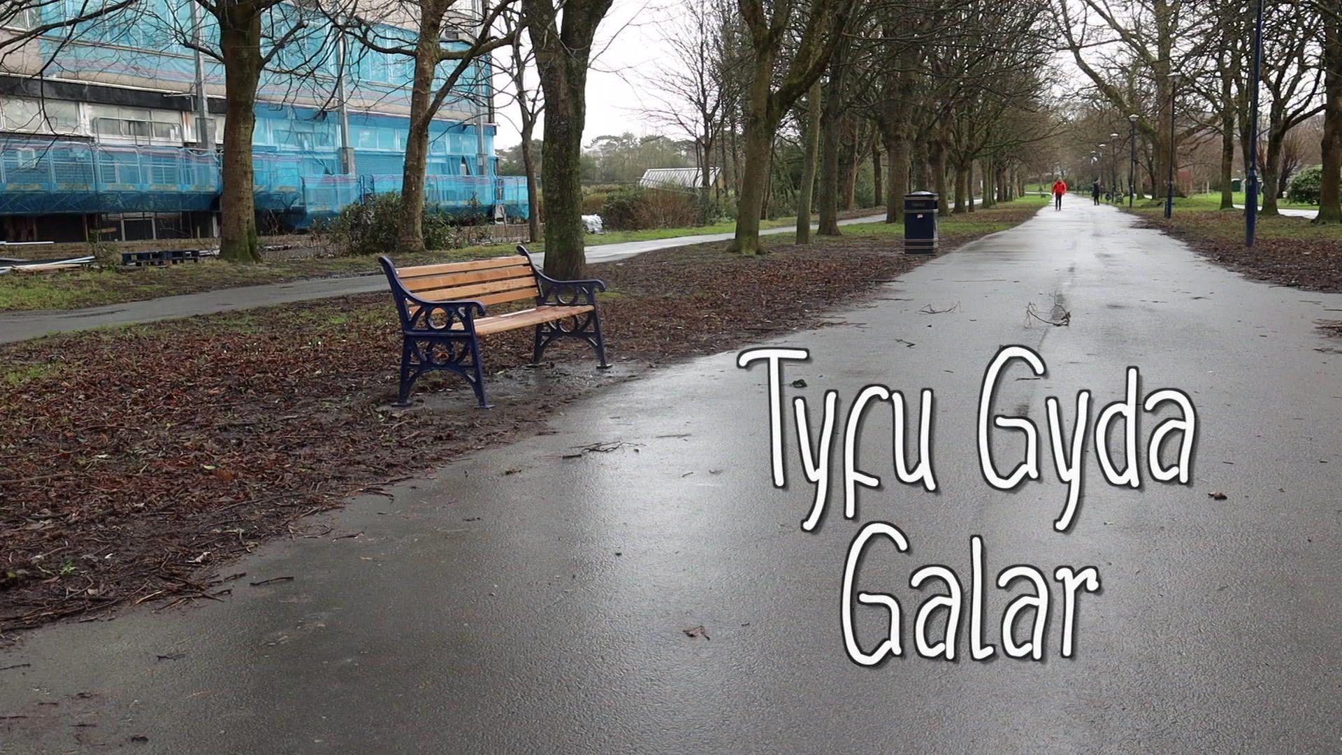 Teitl y ffilm, Tyfu Gyda Galar, dros olygfa ar ddiwrnod hydrefol o lwybr hir gyda choed ar bob ochr. Mae adeilad mawr ar y chwith, a mainc pren ac mae pobl i'w gweld yn cerdded yn y pellter.