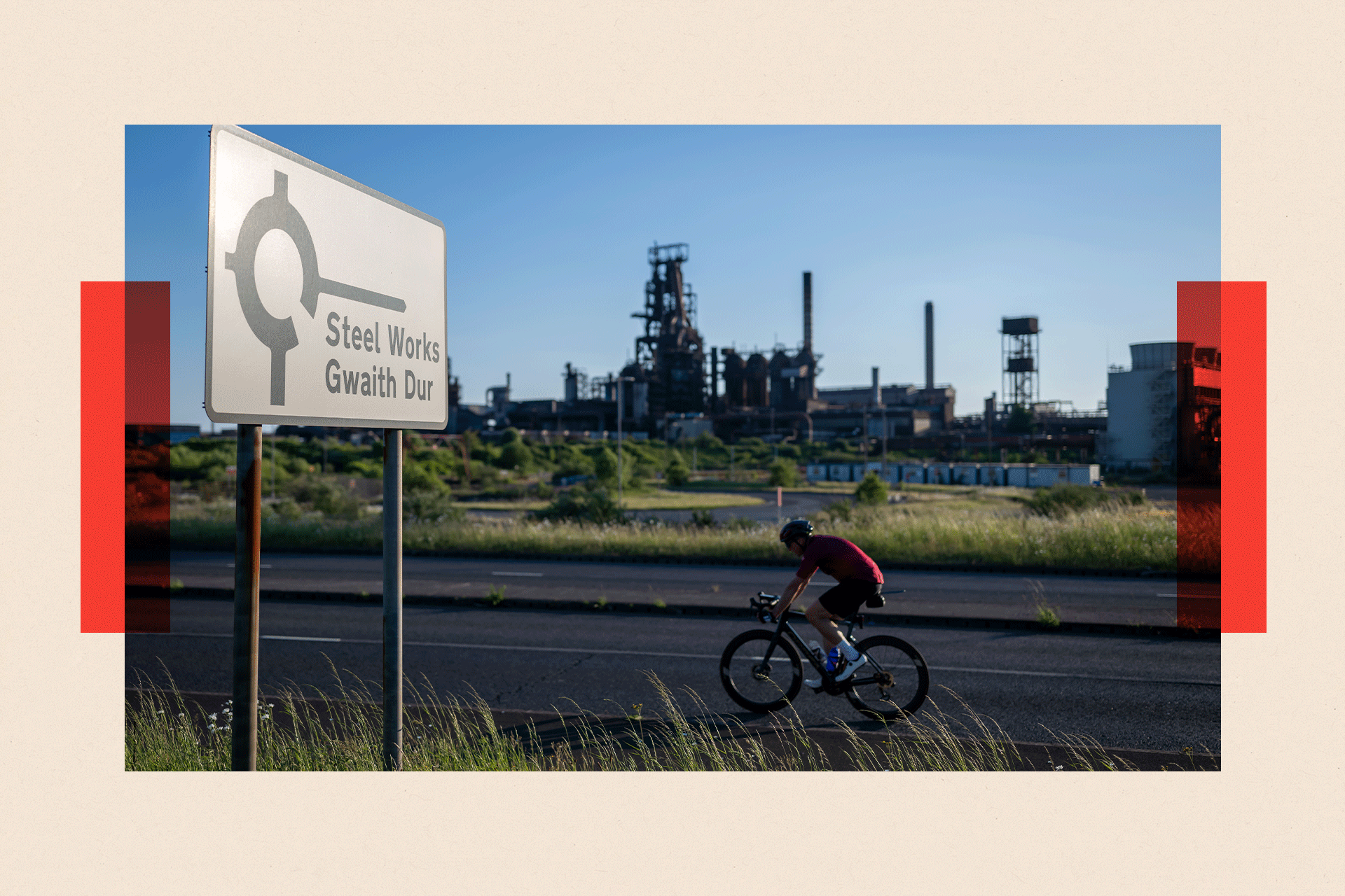 Cyclist in red rides along a road with a sign for the steelworks in the foreground, and the steelworks in the background