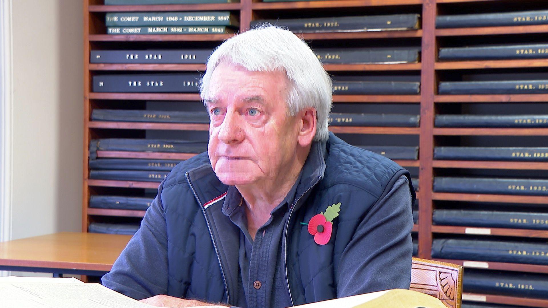 A man with short grey hair in a blue shirt and jacket sits in an archive newspaper room.