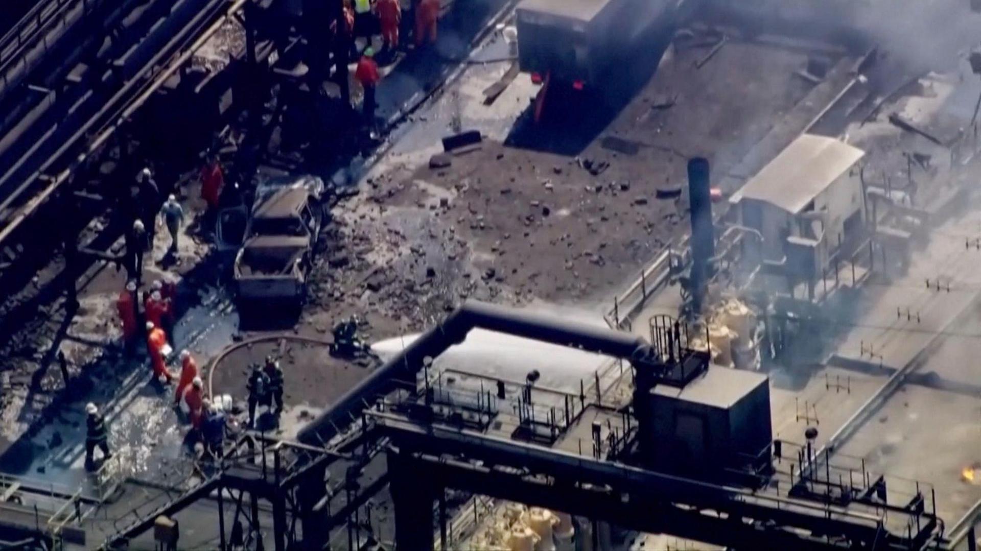 From high above, pipes, metal chimneys and walkways ring an open area with a burned out truck, mild rubble and another paneled truck. People in orange jumpsuits and white hard hats can be seen standing to one side and a fire hose blasts white jets of water.