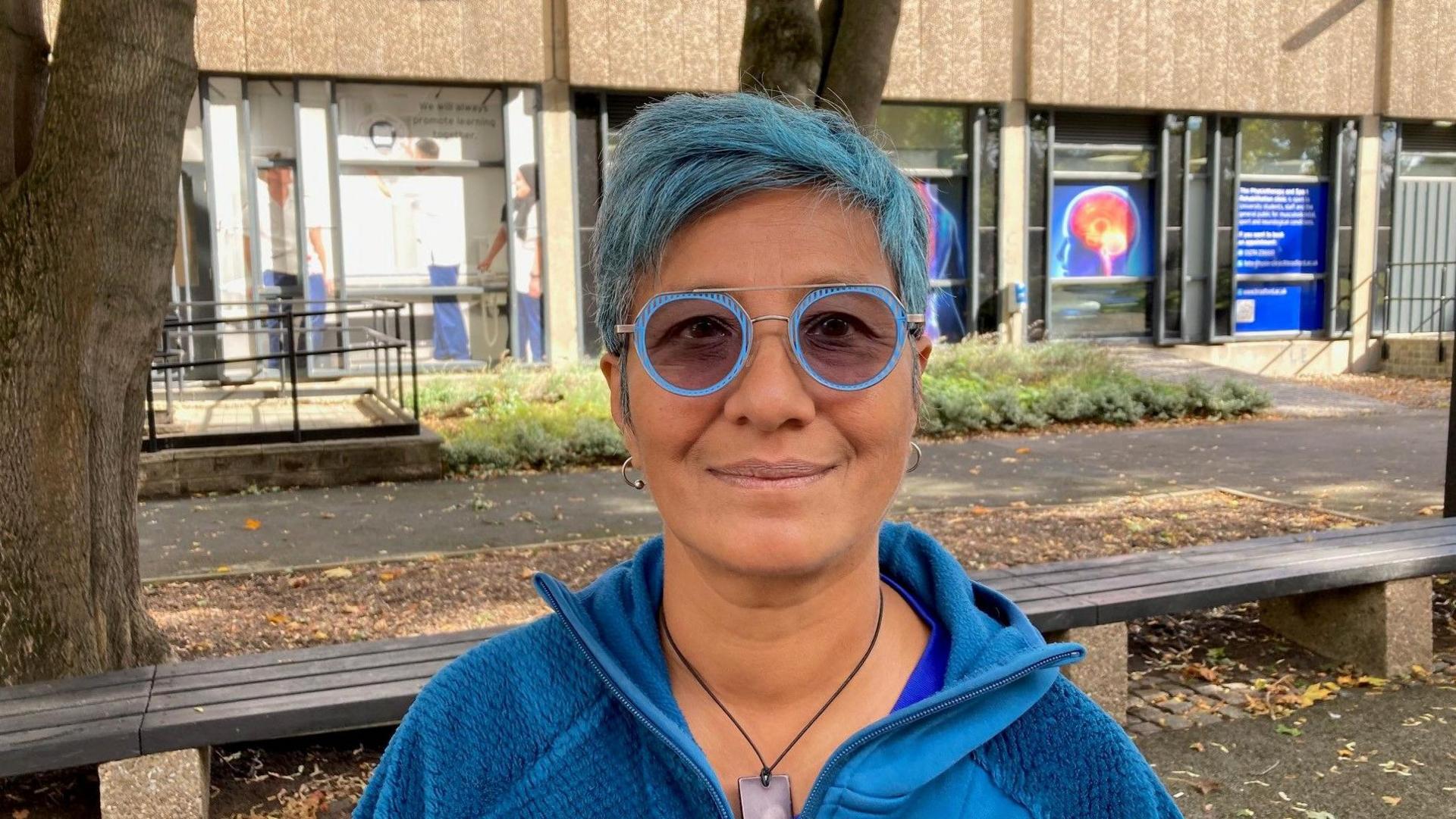 A woman with short, blue-dyed hair wearing light blue-framed glasses and a two-tome blue fleeced jacket. 