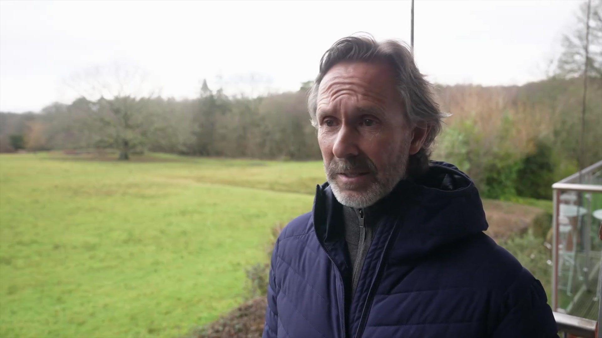 Një burrë qëndron jashtë. Pas tij është një fushë me pemë. Ai ka veshur një xhaketë blu dhe flokë gri të lëshuara prapa.