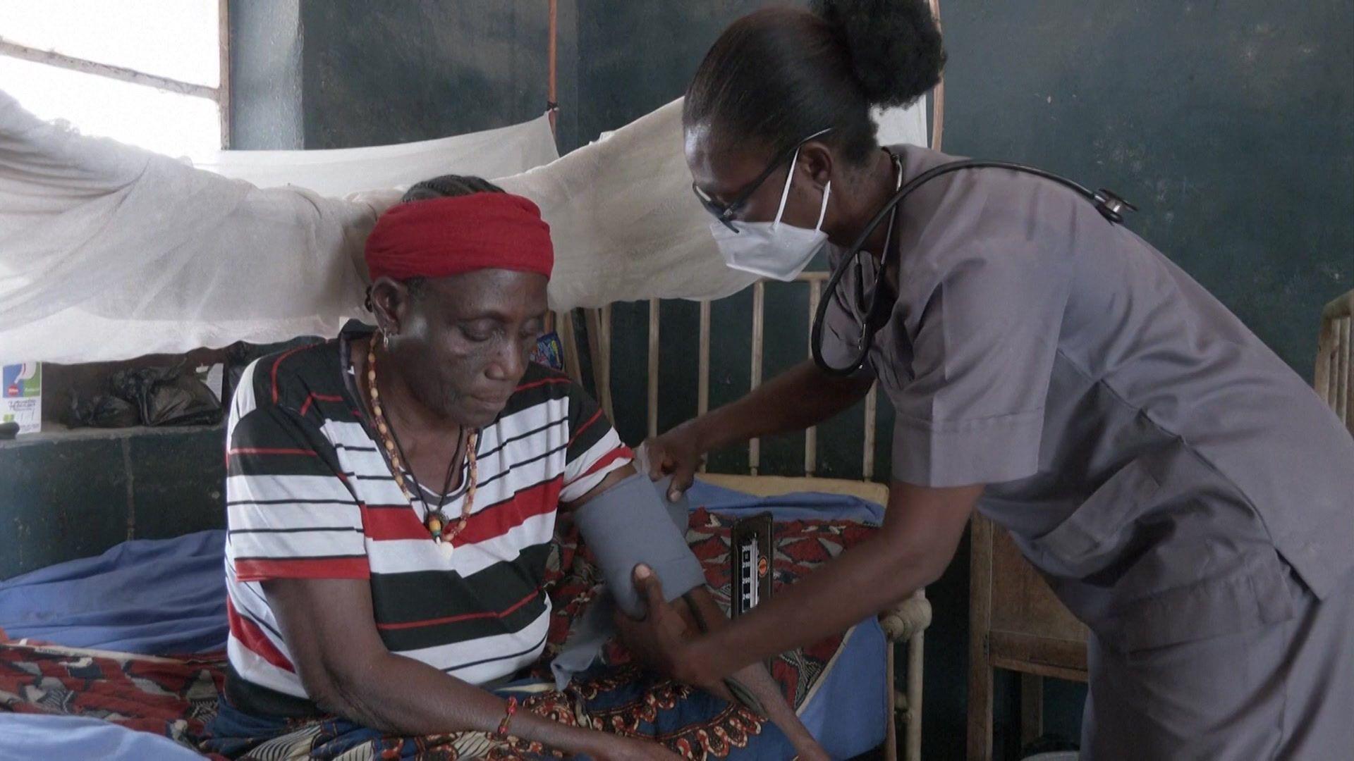 A woman is sitting on a hospital bed while a nurse standing next to her is taking her blood pressure.