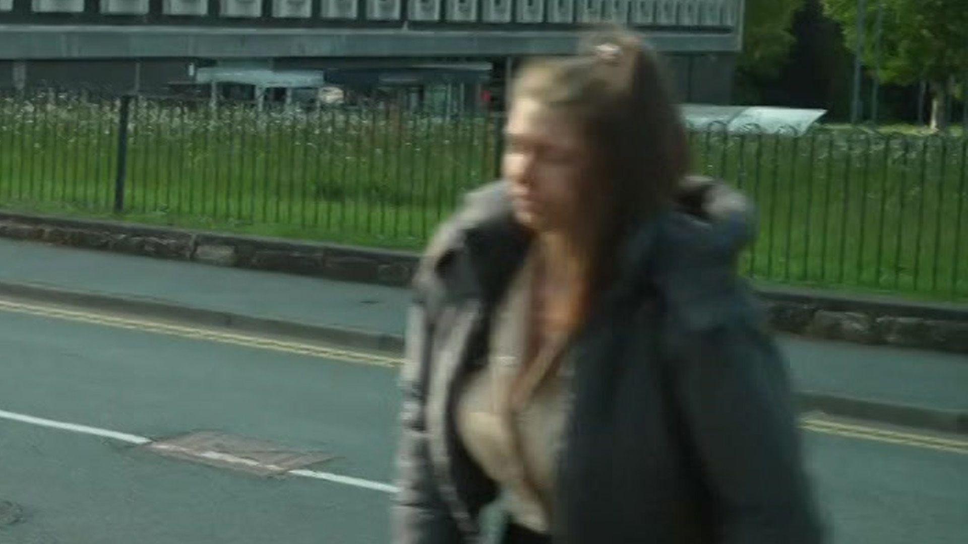 A blurred photo of a woman walking across a road. She has her brown long hair tied up in a ponytail. she is wearing a grey coat.