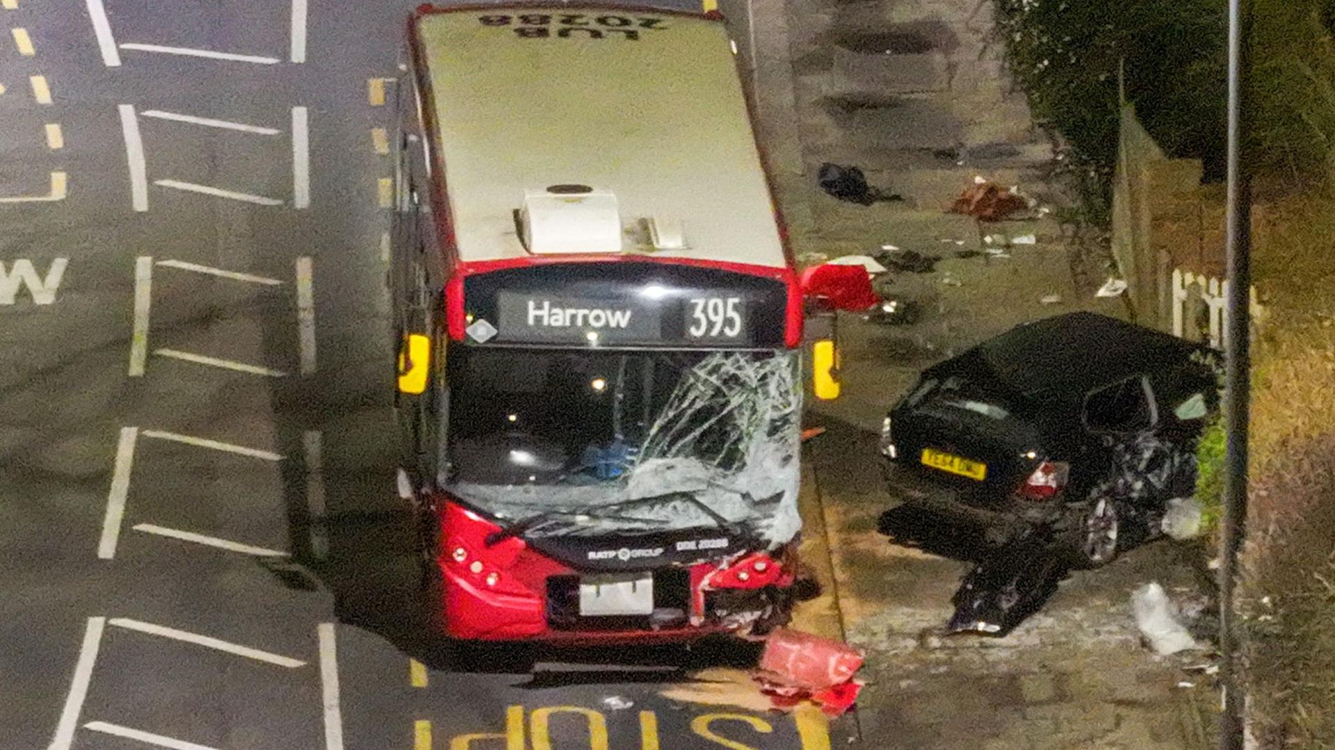 395 bus windscreen smashed and a black vehicle to its right crashed into a fence with debris from both cars strewn across the bus stop