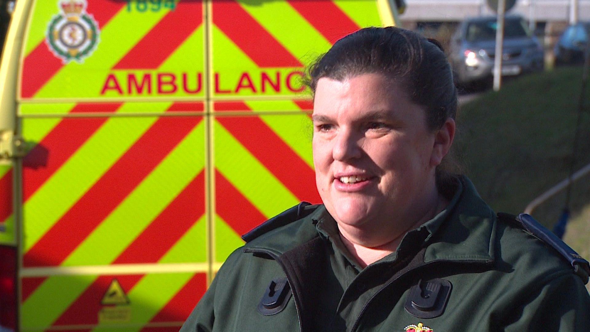 A woman with brown hair tied back wearing dark green ambulance attire standing in front of an ambulance van.