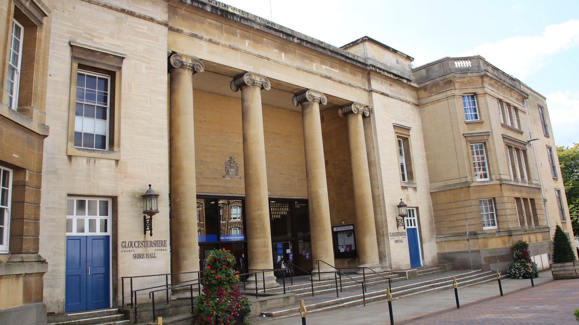 The front exterior of Shire Hall in Gloucester on a sunny day. It is a grade II listed building in Greek revival style, with large columns at the entrance and blue doors on either side.