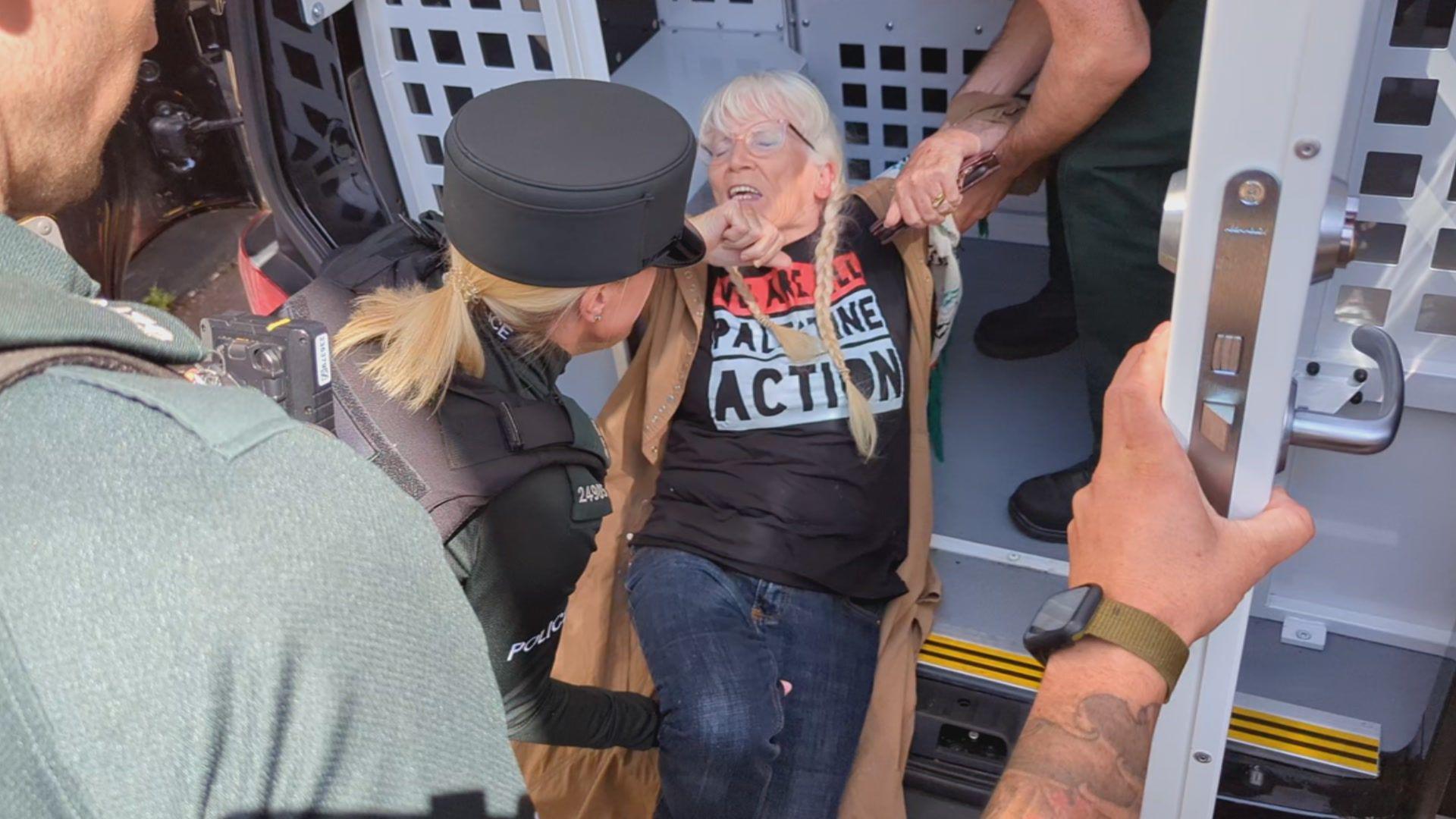 A woman with white hair being moved into the back of a police van. Two police officers are next to her.