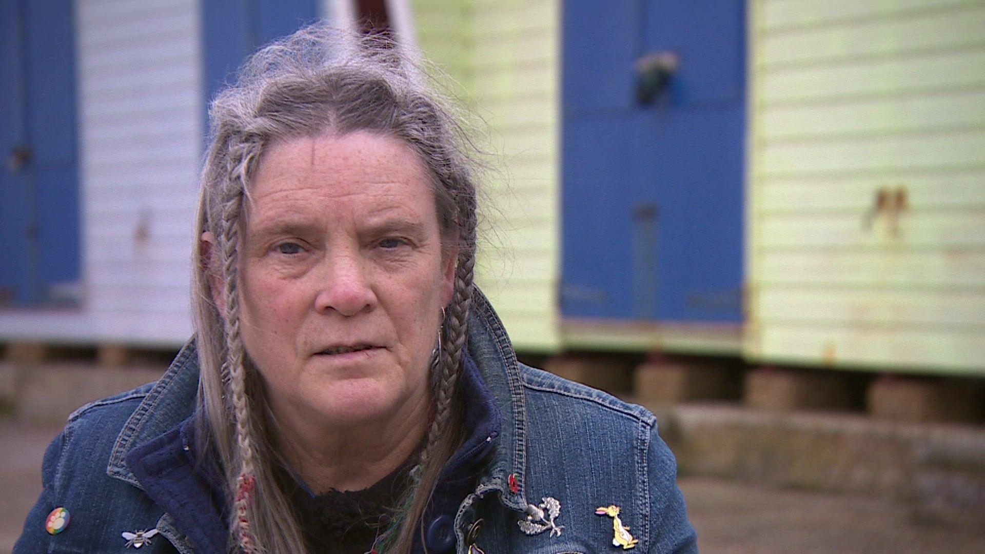 Phazar Souter. She has platted grey hair and is wearing a denim jacket with various pins on it. Out of focus behind her are colourful beach huts.