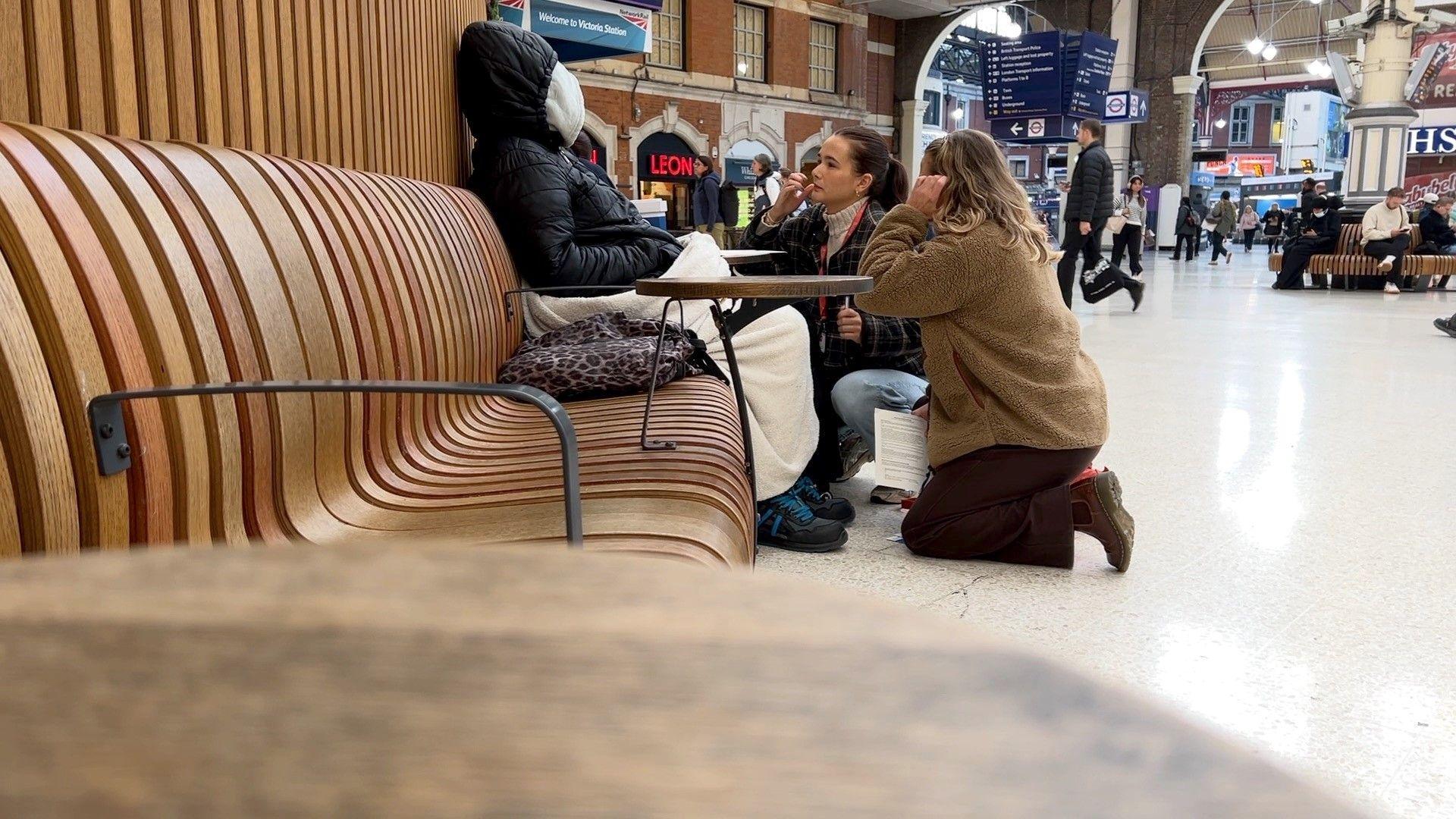 Two young women kneel to talk to a woman wrapped in a blanket and hood jacket at a train station
