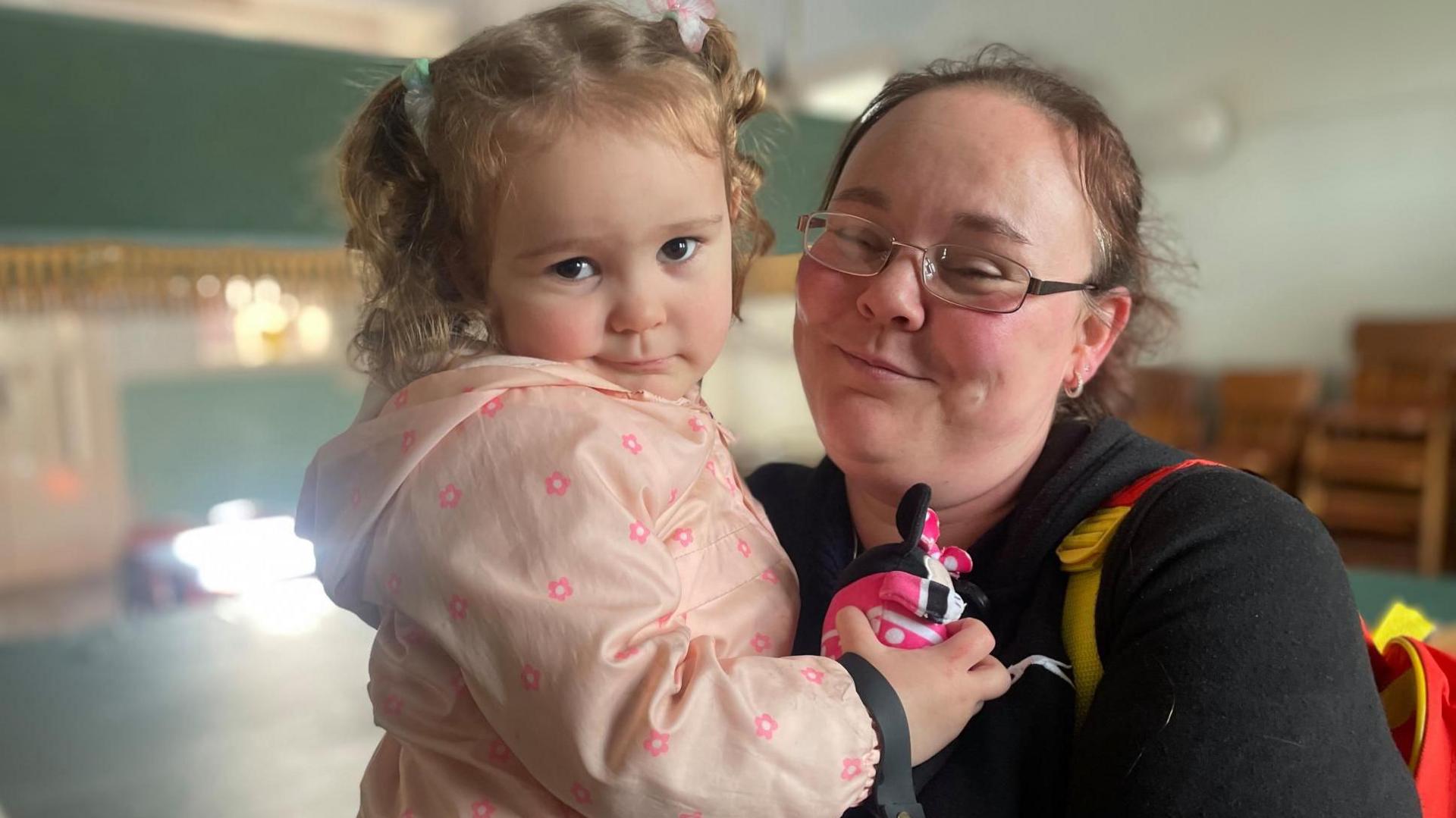 A woman with hair tied back holds a small toddler in bunches clutching a pink toy.