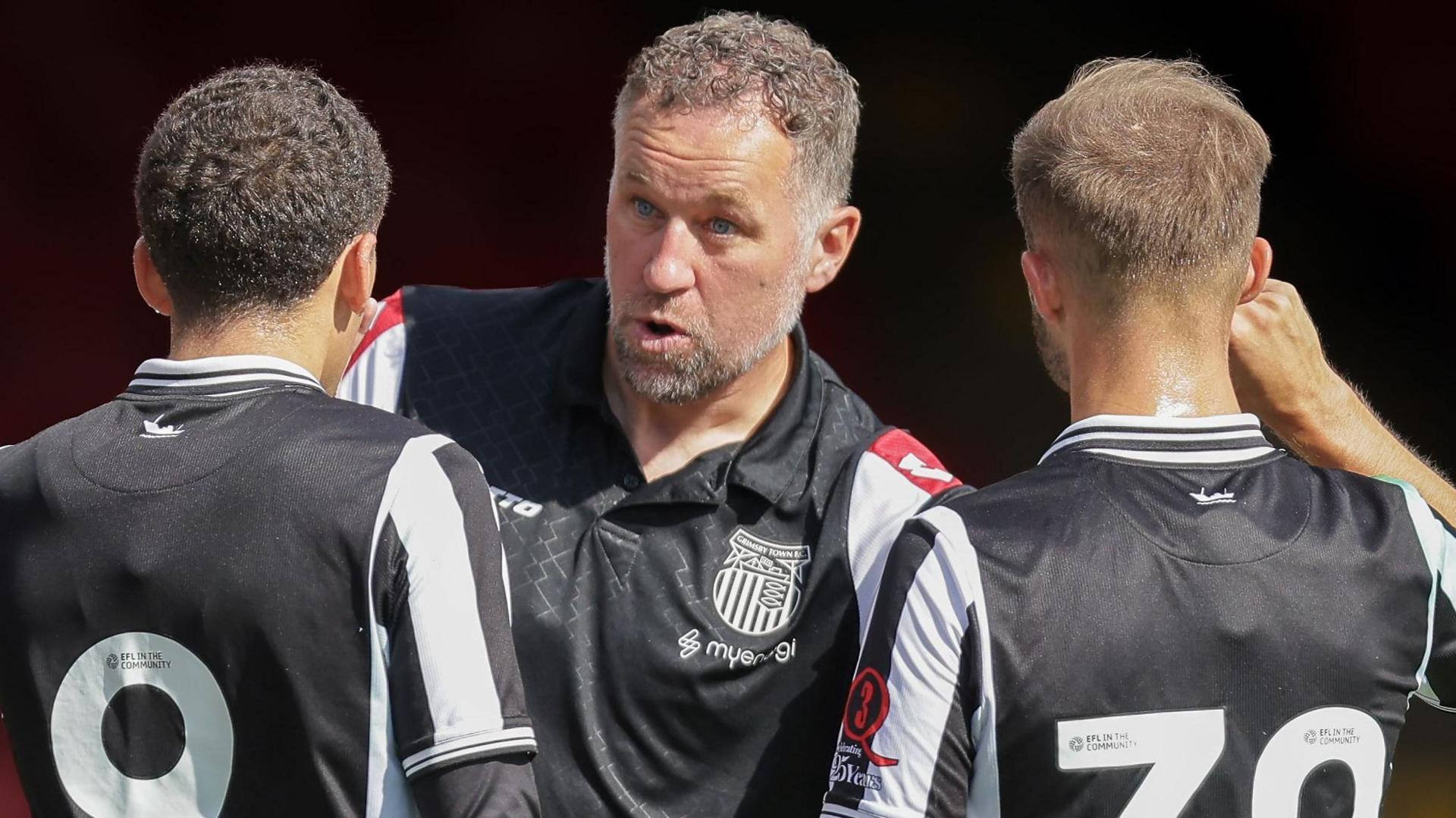 David Artell, wearing a black Grimsby Town training top with white and red patterened sleeves, speaks to his players during a pre-season friendly