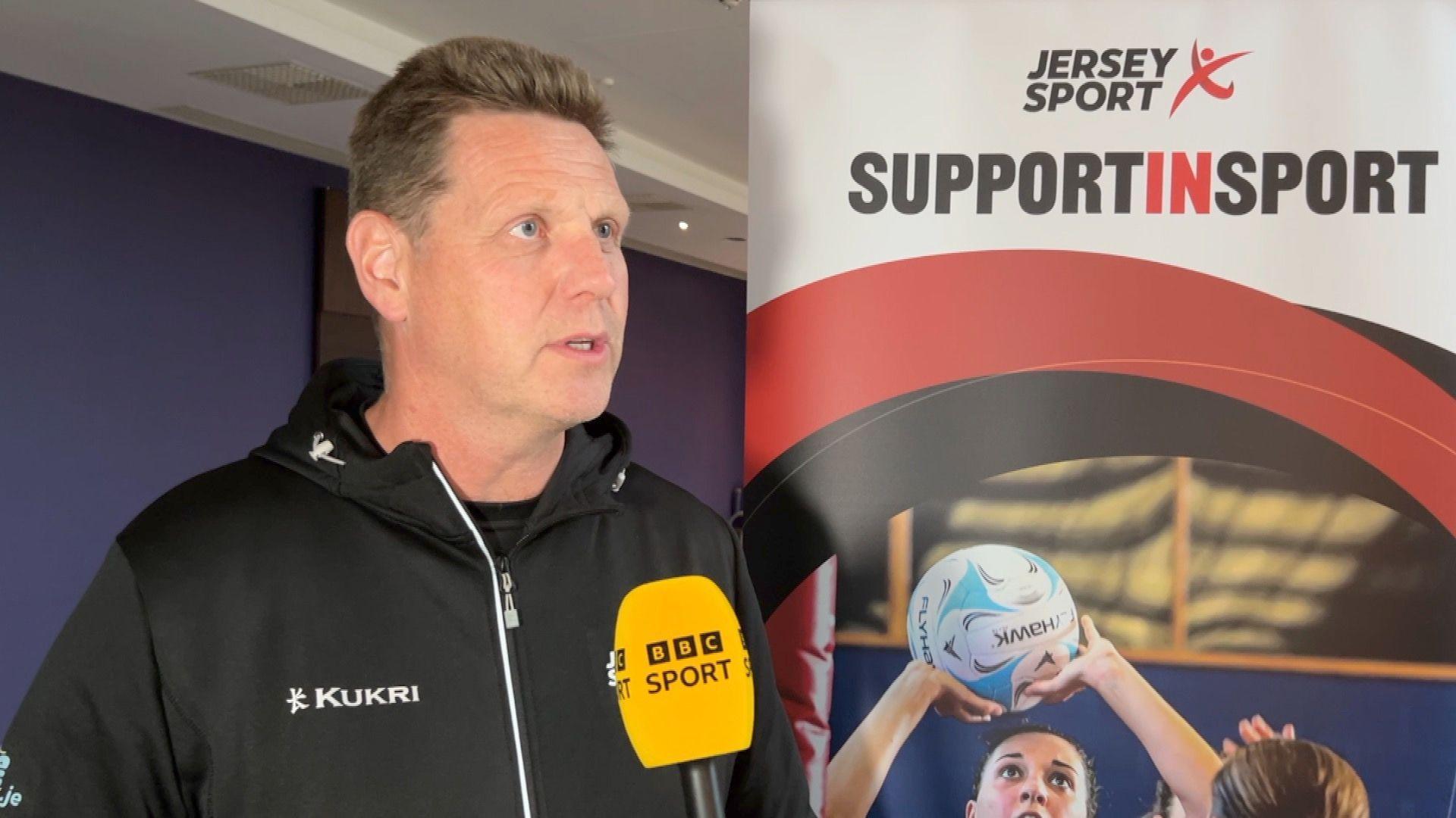 David Kennedy, a man with brown hair, wearing a black zip-up hooded jumper. He is standing on the left, looking right, with a yellow BBC Sport microphone in front of him. Behind him is a sign which says: "Jersey Sport, support in sport." It includes an image of a woman playing netball.