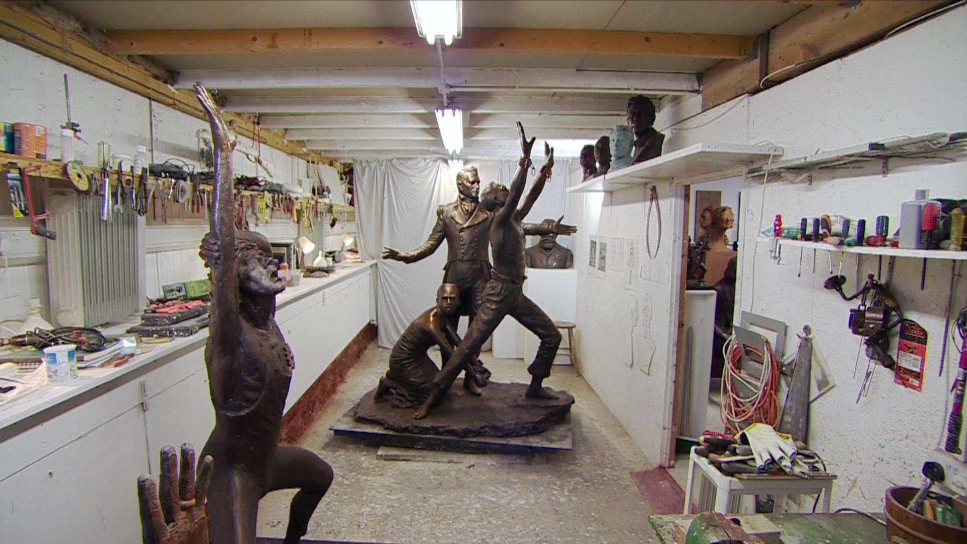 A bronze sculpture in a workshop. The walls are white and you can see tools hanging and drawings on them. The sculpture shows three figures – a shackled female slave kneeing down, an officer from the West Africa Squadron and a freed slave with his arms stretched to the sky. In the foreground there is a second sculpture of a standing man who who appears to be in pain.