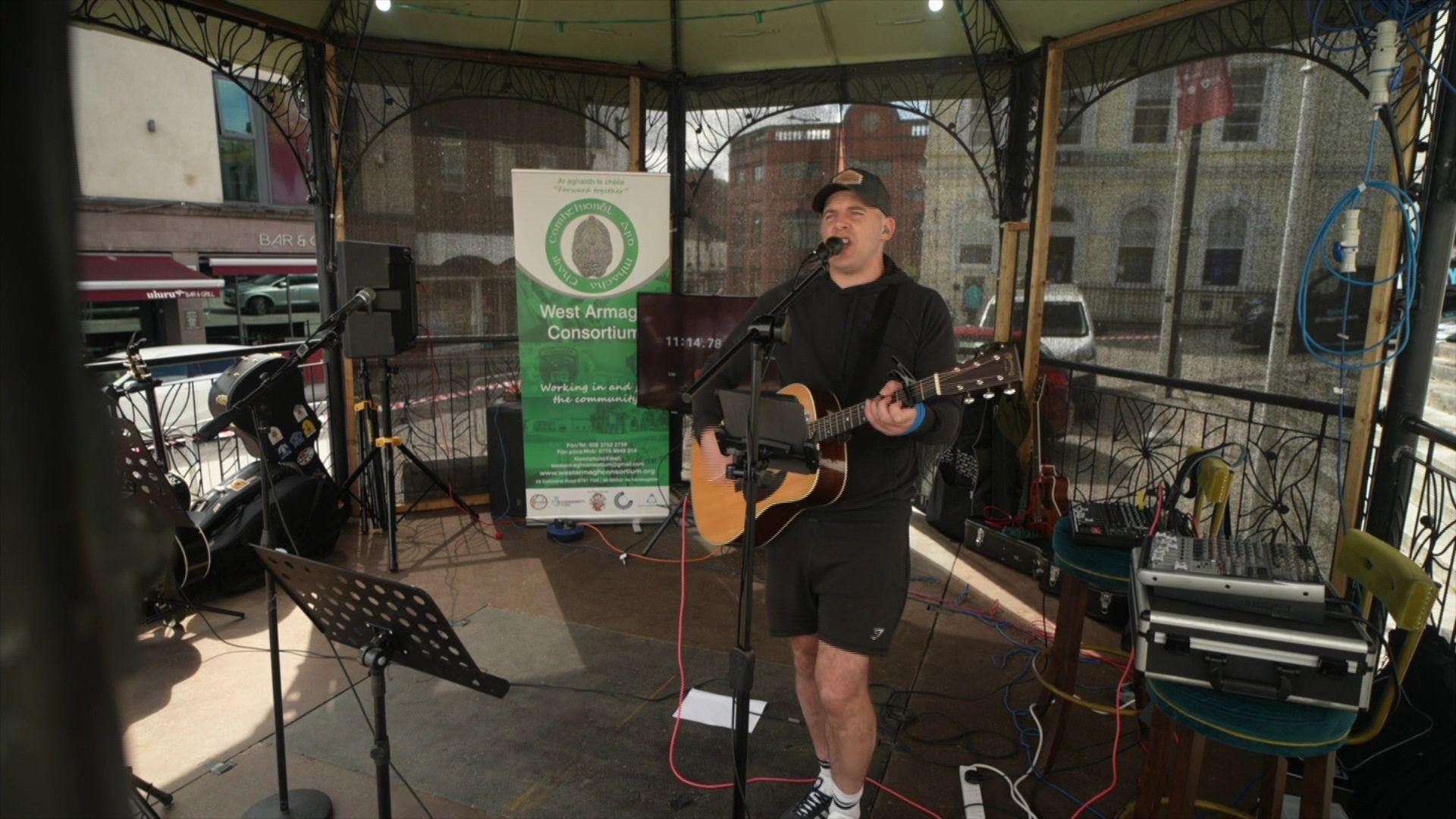 Martin Rafferty plays his guitar under a metal gazebo. He wears a black hoodie and a baseball cap. He has blue tape around his hand.