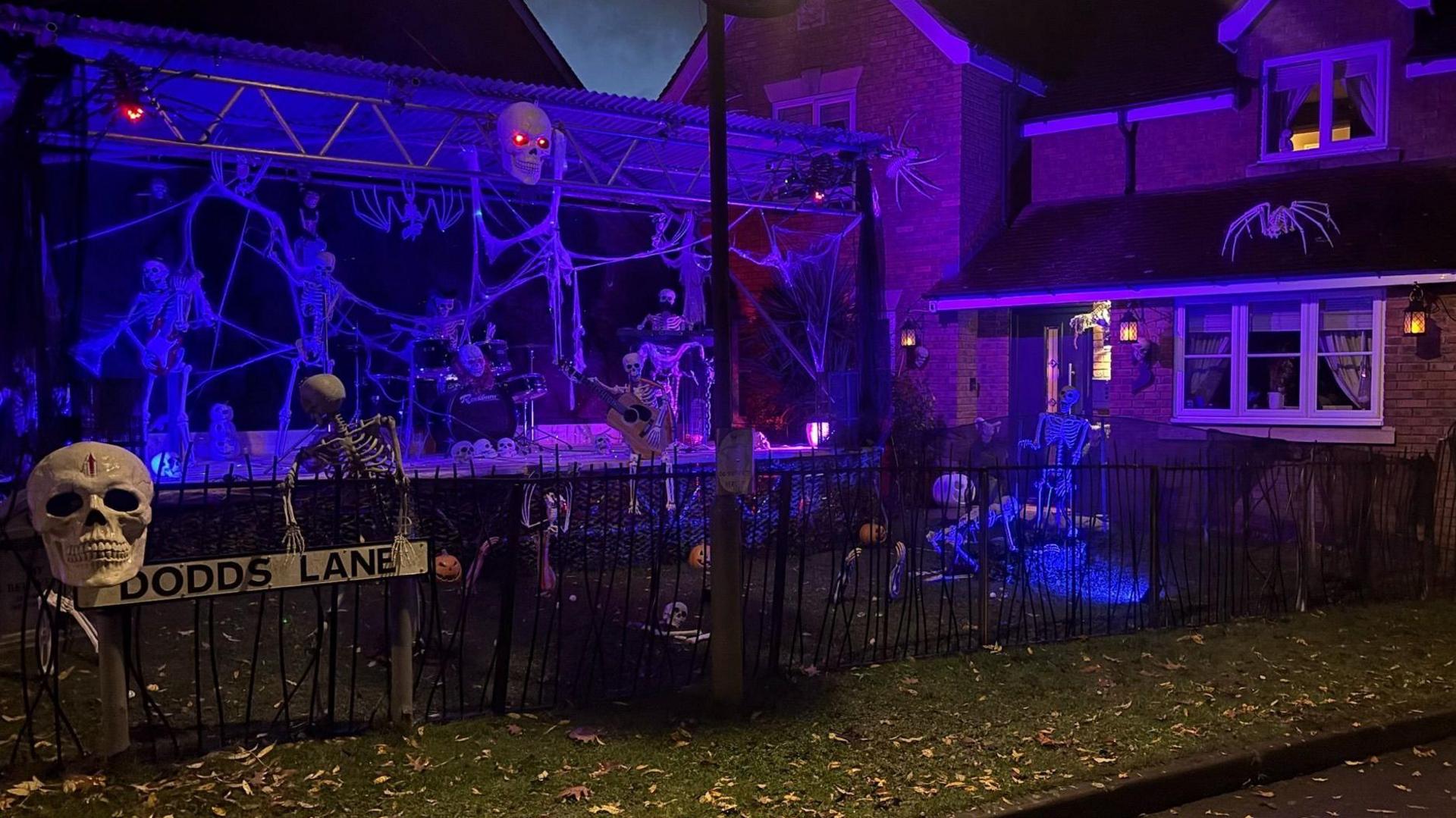 A stage with a skeleton playing on a drum kit and others playing guitars. There is a house on the right hand side of the image and cobwebs hanging around the stage. A sign on the left hand side of the image reads Dodds Lane.