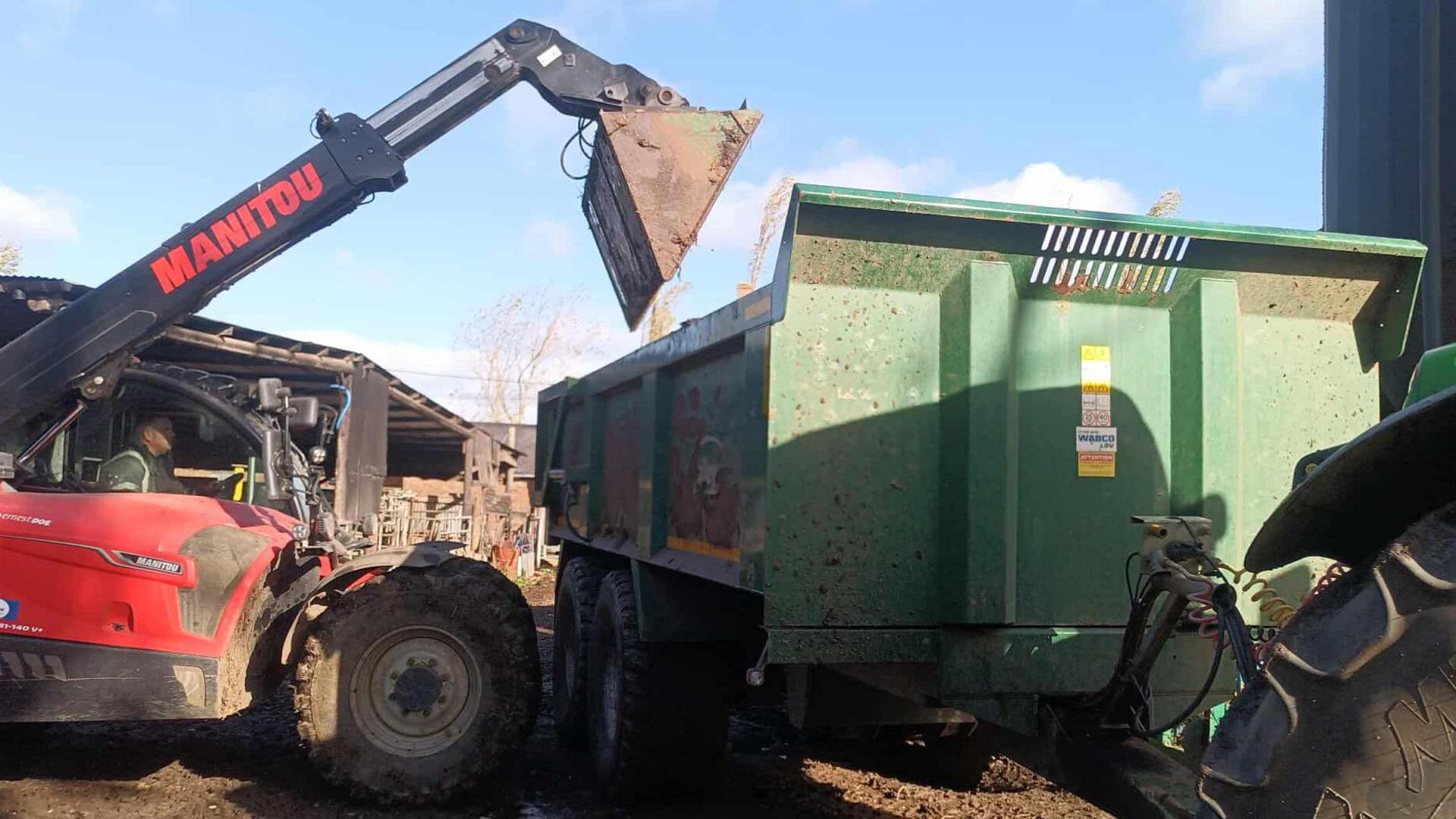 A green farm trailer is being loaded with manure by a red tipper with an extendable black arm with a large bucket on the front, farm buildings and barns can be seen behind.