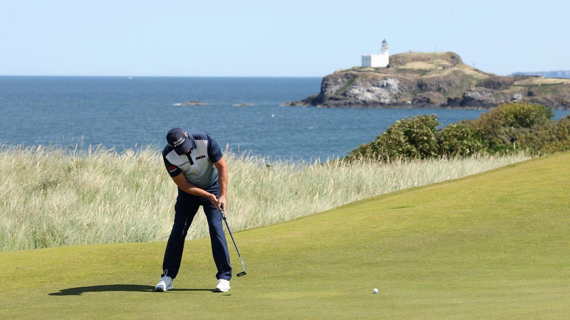 Padraig Harrington rolls a putt on the 13th green during the pro-am. He is wearing a navy cap, white and navy polo shirt, navy trousers and white golf shoes. A white lighthouse is visible on a rock in the background.