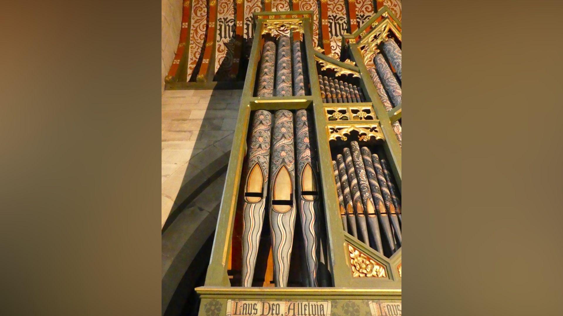 Green-painted casing around black and gold pipework on a 19th Century organ in a church. Just the top of the pipes can be seen, stretching up to a red and black painted ceiling.