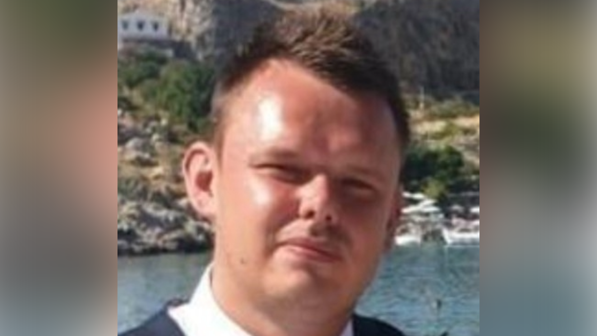 A man with brown hair standing in front of a lake in the sun
