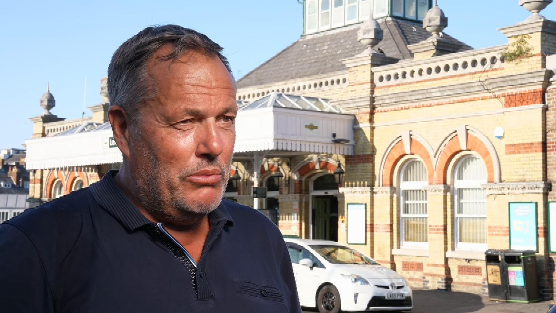A man in a black collared shirt is standing outside a train station. He has greying hair. There are cars parked outside the station.