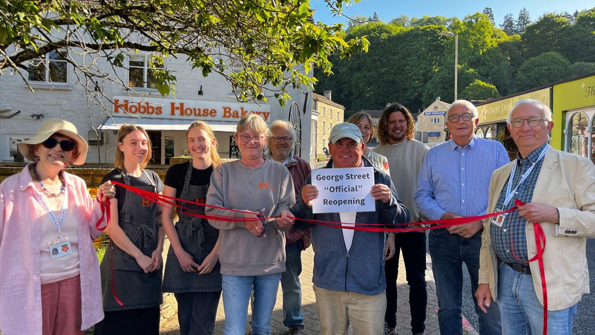 A group of people holding a paper sign with a red ribbon being cut in front of a bakery in Nailsworth