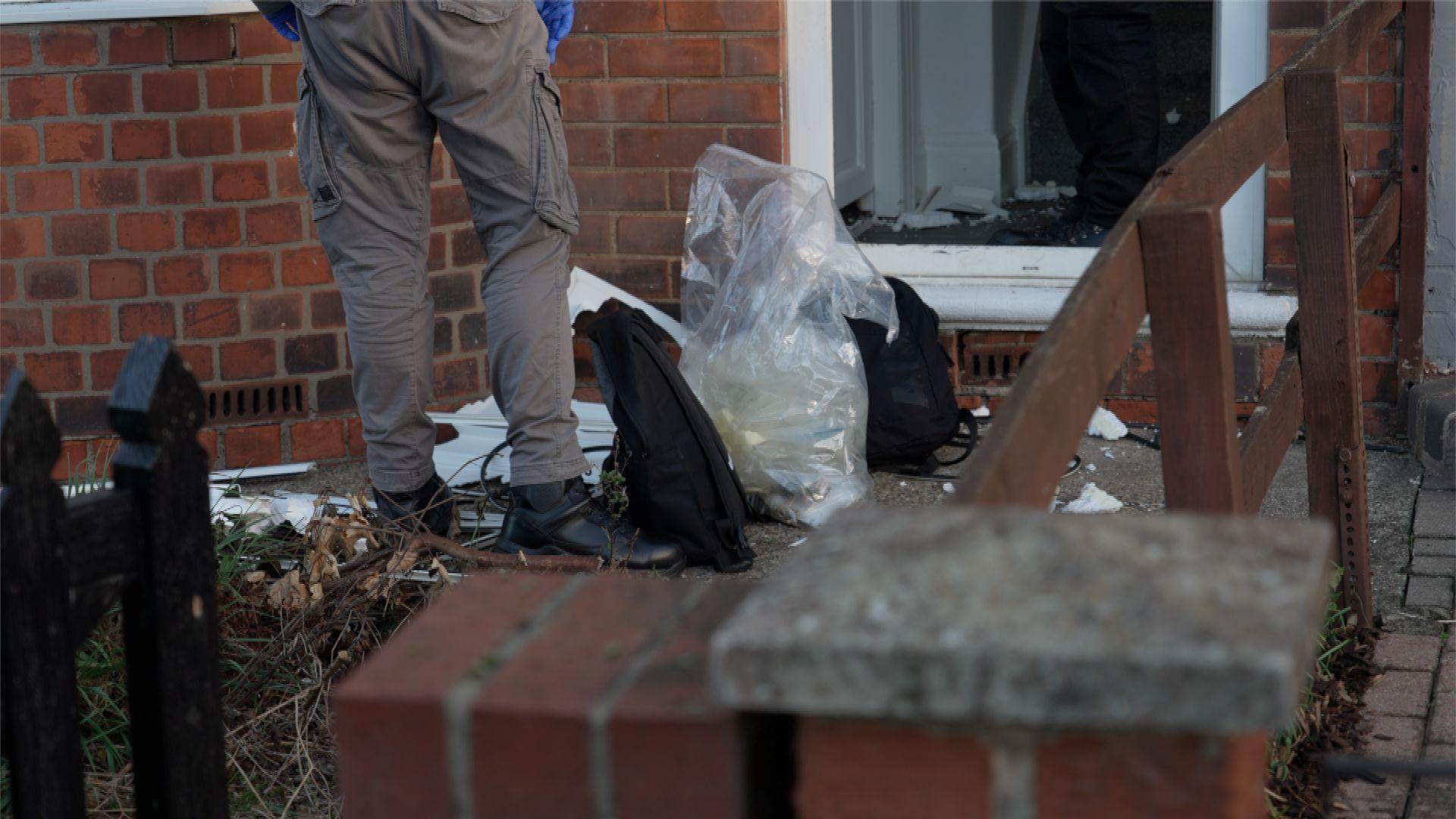 A police officer stands next to two black bags and a large clear sack which have been places on the path of a house as it is searched by officers. The door of the property has been smashed and pieces are strewn on the floor.
