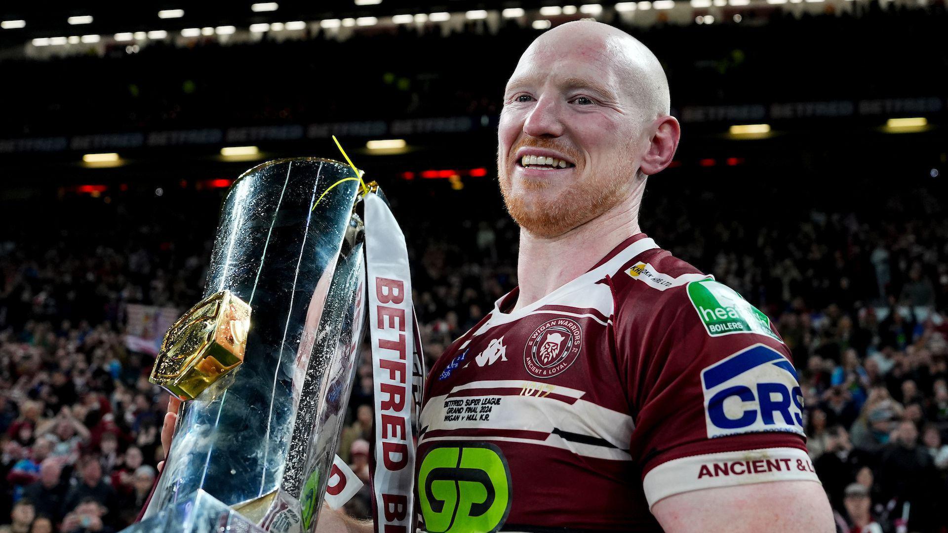 Liam Farrell smiles toward the camera as he clutches the Super League trophy on the pitch at Old Trafford in the aftermath of the win over Hull KR in 2024