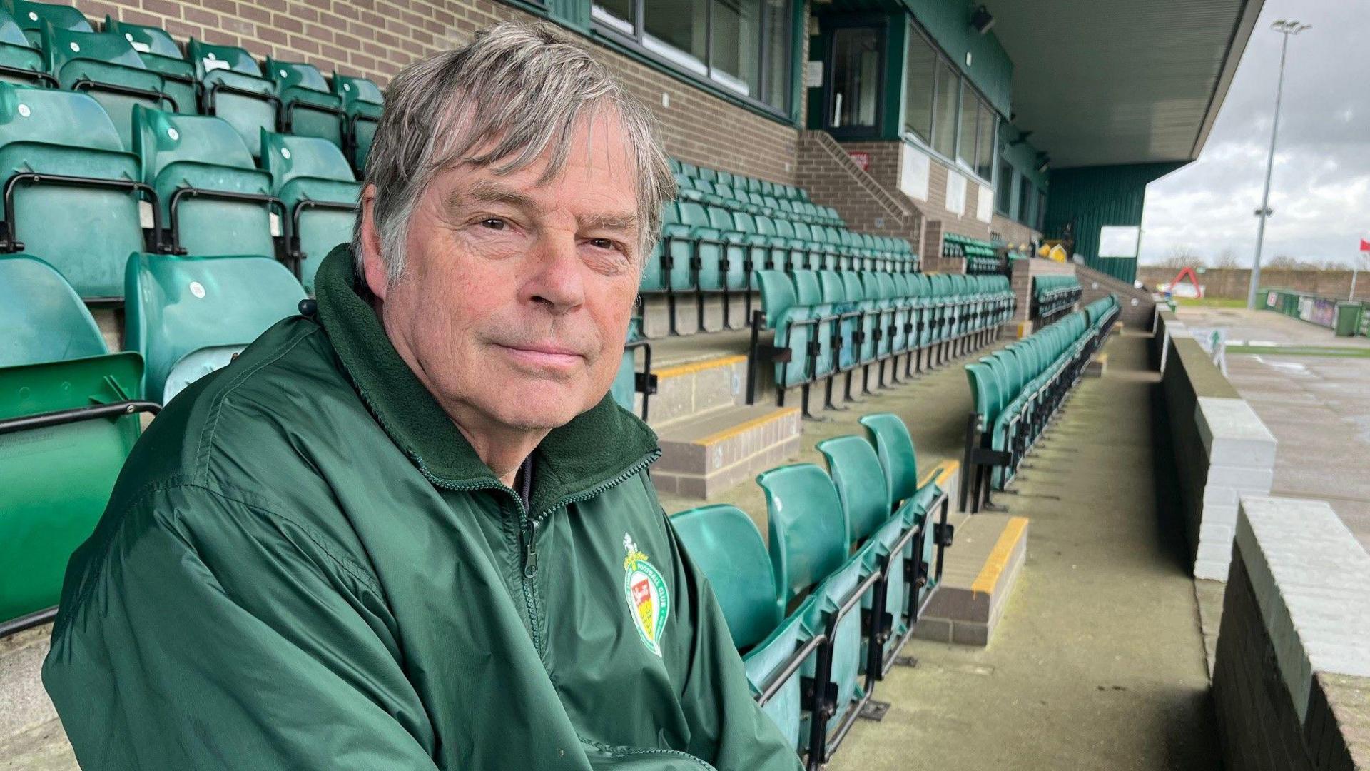 A man wearing a green coat sitting in green seats in a football stadium.