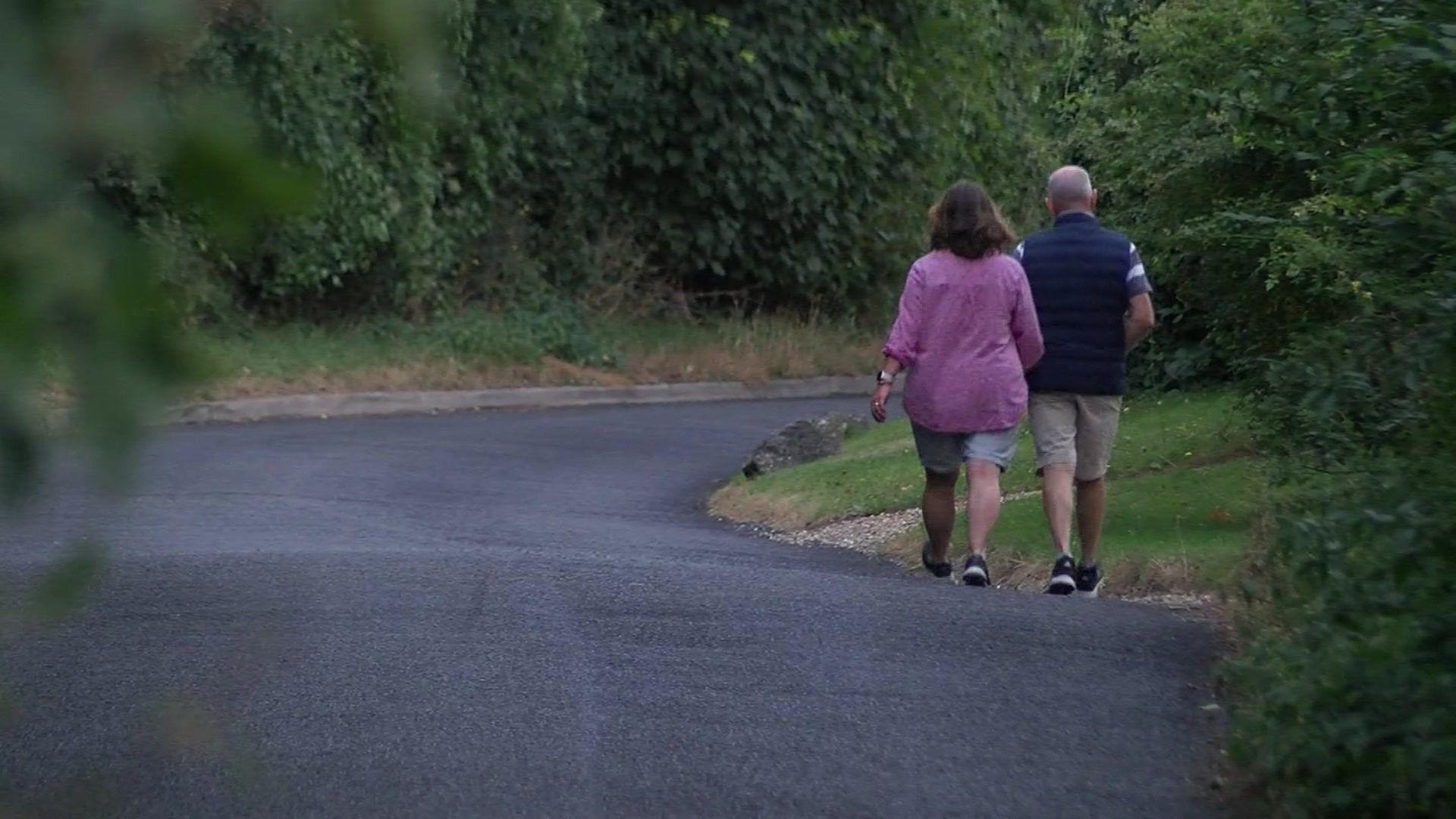 A woman wearing a pink top and light grey shorts and a man wearing a gilet and beige shorts walk along the edge of a road with their backs to the camera. There's no pavement so they're having to walk in the road and they're approaching a blind bend.