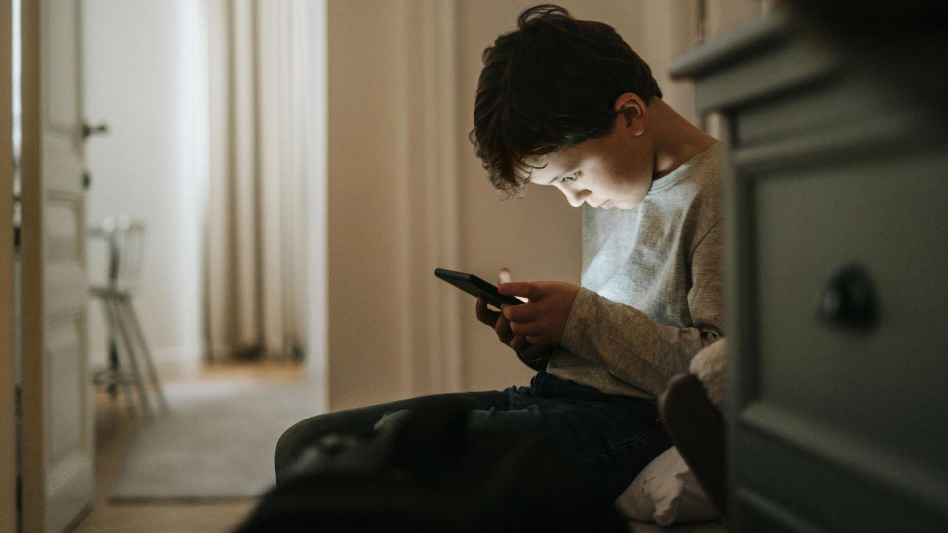 A boy sits on the floor looking at his smartphone.