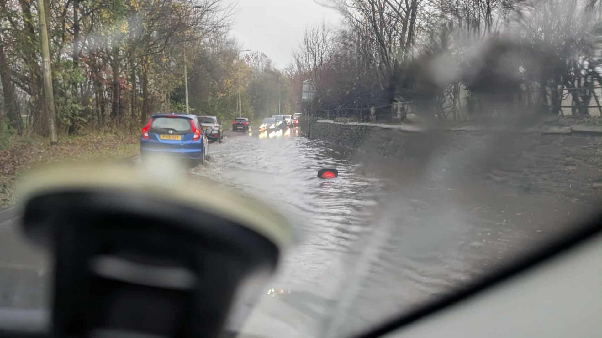 A picture taken from inside a car looking out at a line of cars driving on the extreme left-hand side of the road to escape the road surface flooding in Great Corby.