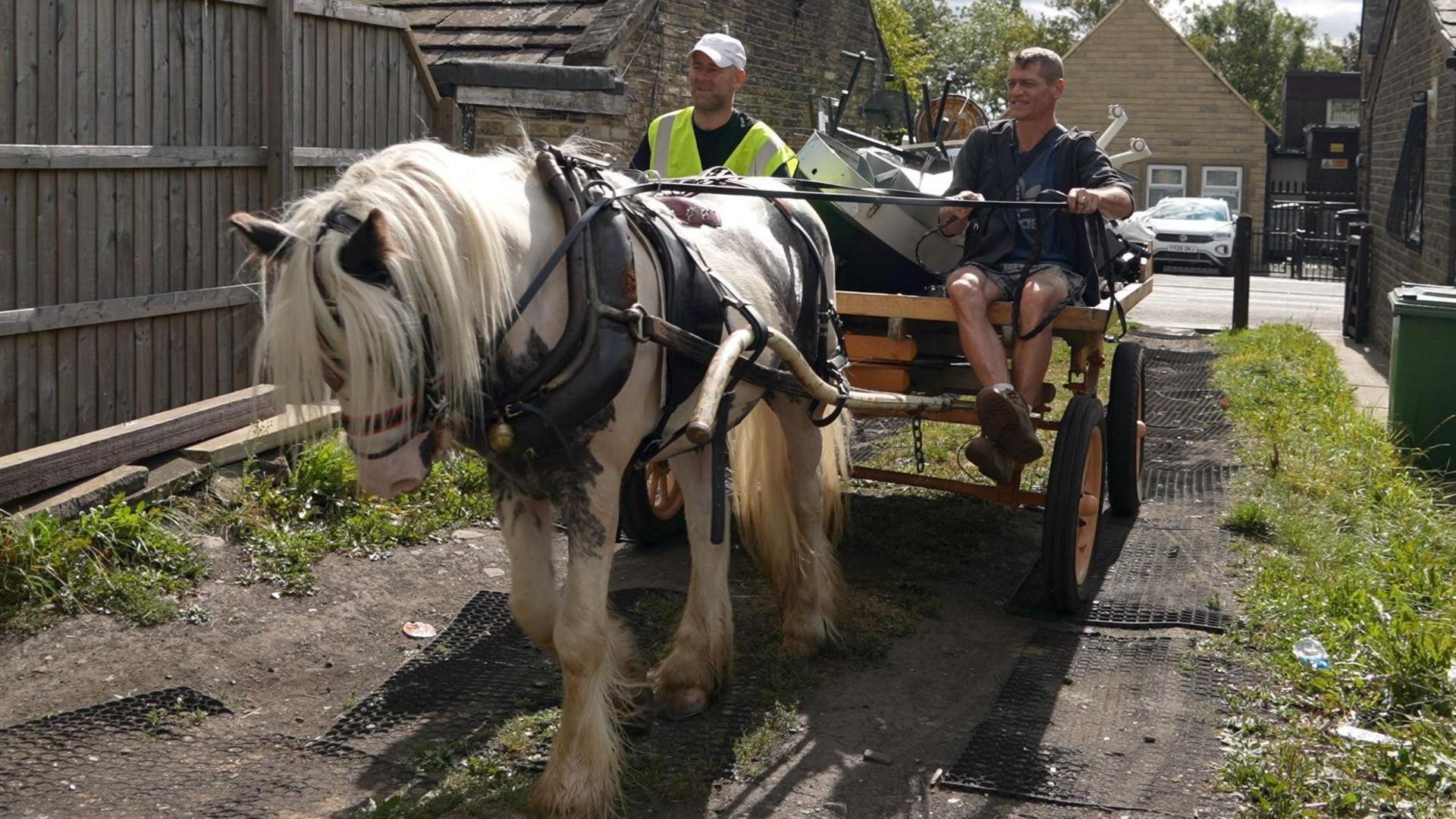 Two men sat on a horse-drawn cart which is being pulled through a track off a street. The cart is piled with what appears to be rubbish.