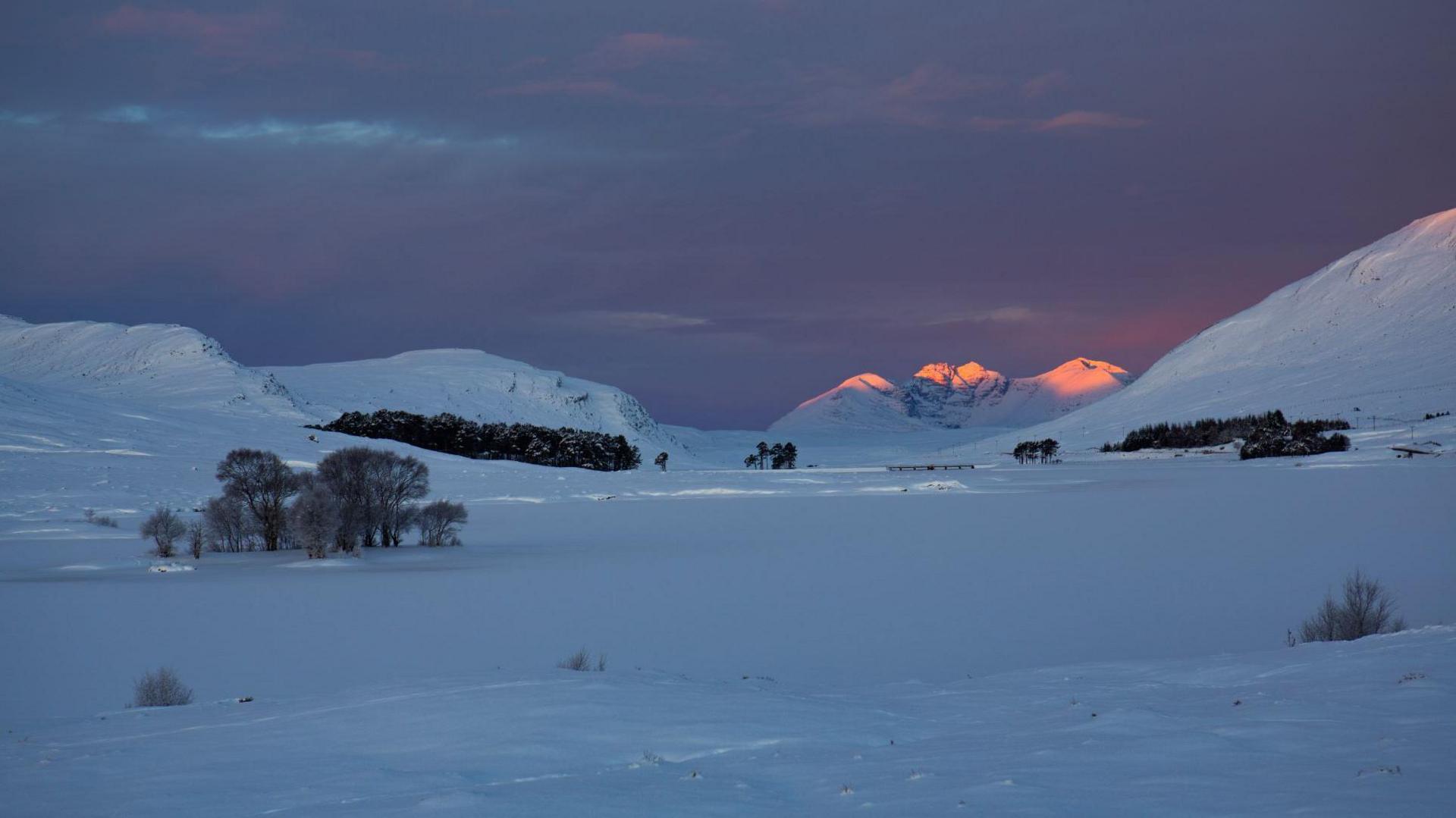 Snow covers a Scottish Highland landscape. The frozen loch stretches to mountains including An Teallach, which is catching sunlight.