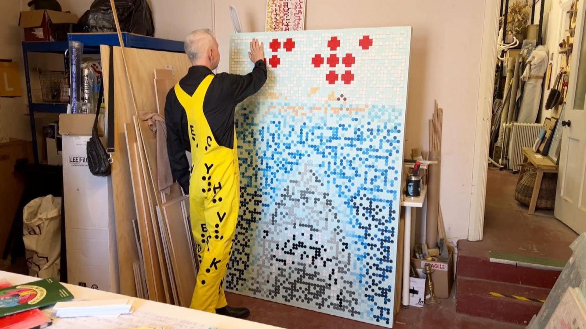Mr Reynolds is again in his studio, wearing the same outfit. He has his back to the camera and is facing his artwork, called 'Jaws' which is a braille imitiation of the original movie poster. Mr Reynolds is holding his hand up to the top left corner of the artwork to read the braille. The poster shows red dots, which spell out Jaws in braille. The outline of a swimmer in the water and the head of a shark emerging from the bottom of the poster.