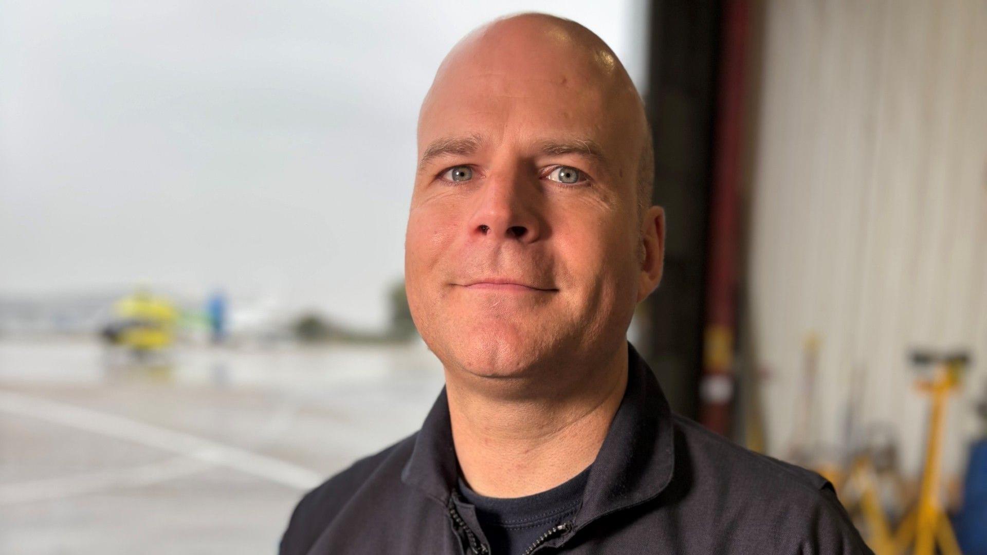 A headshot of Pete Winn, a blue-eyed man who is smiling at the camera. He is wearing a blue jacket and has a bald head. An out-of-focus helicopter is visible in the background. 