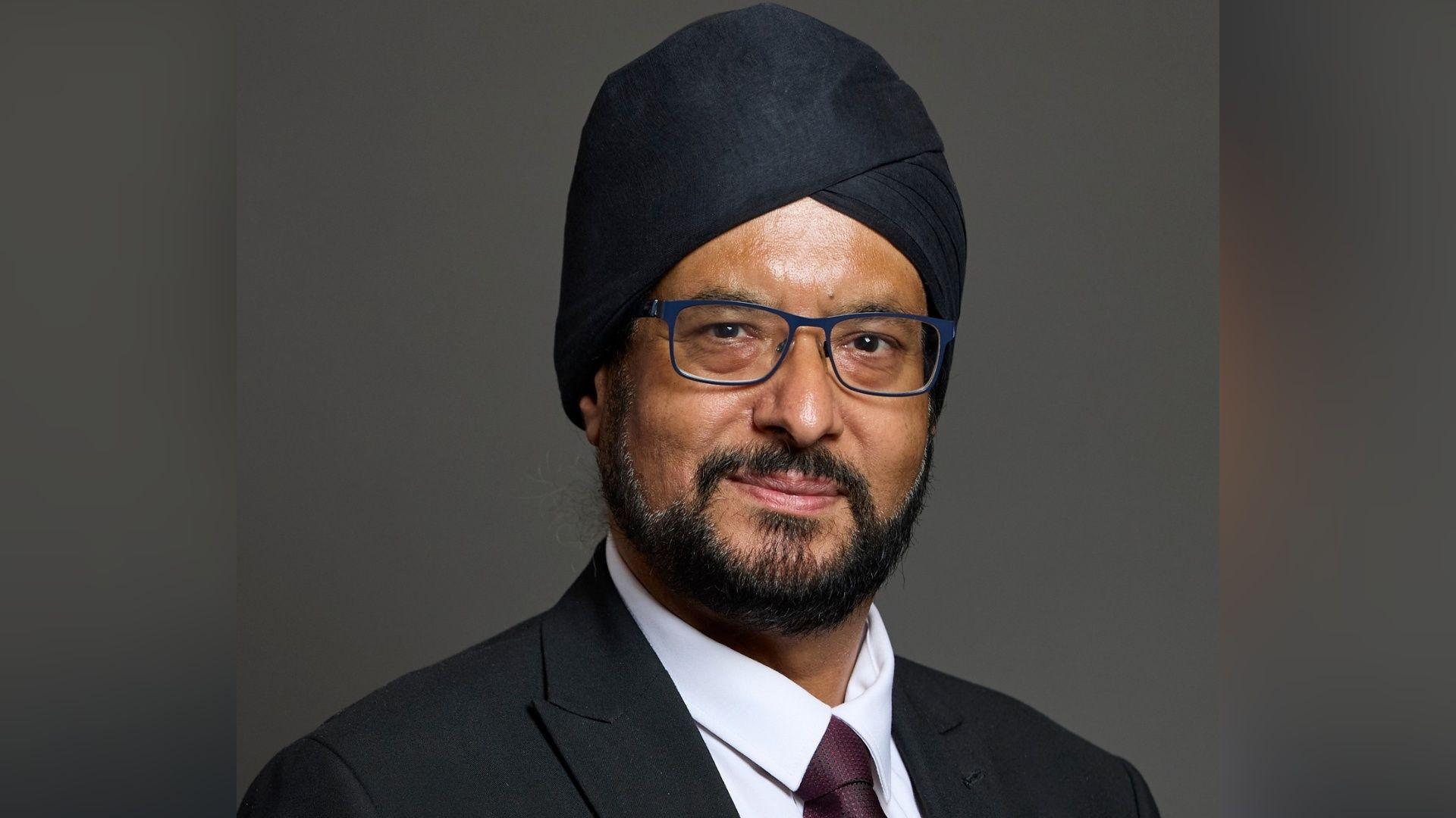 Warinder Juss has a short black beard and moustache and blue glasses. He is looking at the camera with a slight closed mouth smile. He is wearing a black turban, black blazer, white shirt and brown tie.