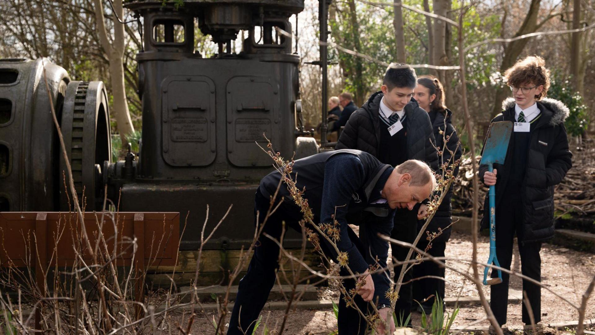 A man with a spade digging the ground. Behind him are parts of old locomotive trains. School-aged teenagers watch the Prince digging. Trees surround the garden and it is winter so the trees are bare of leaves.