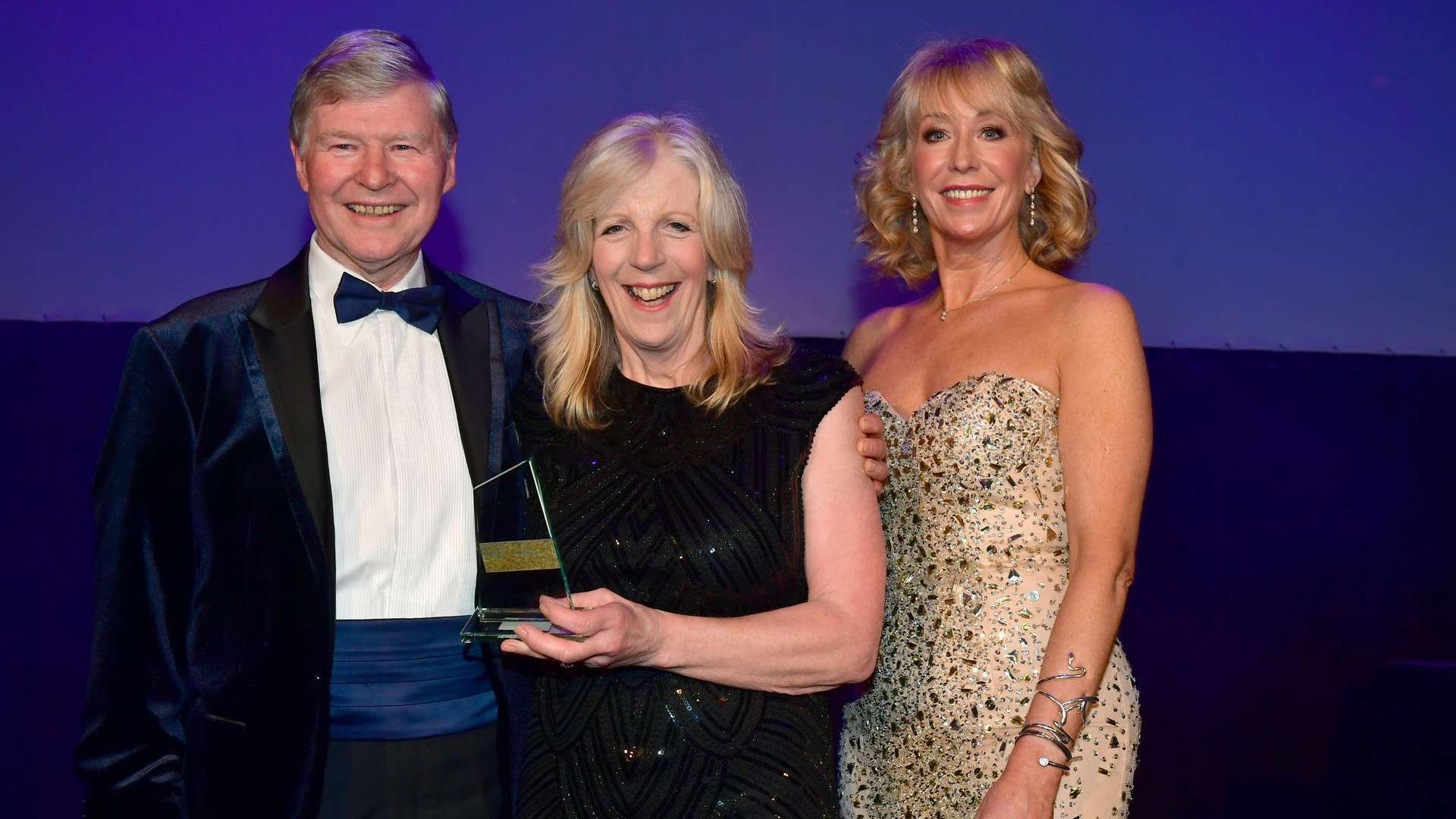 Geoffrey Cox, Margot Whittaker and Emma Jesson stood in a row at the awards. Mr Cox is wearing a tux. Ms Whittaker has blonde hair, she is wearing a sparkly black dress and is holding an award. Ms Jesson has is stood on the left. She has blonde hair and is wearing a sparkly gold dress.