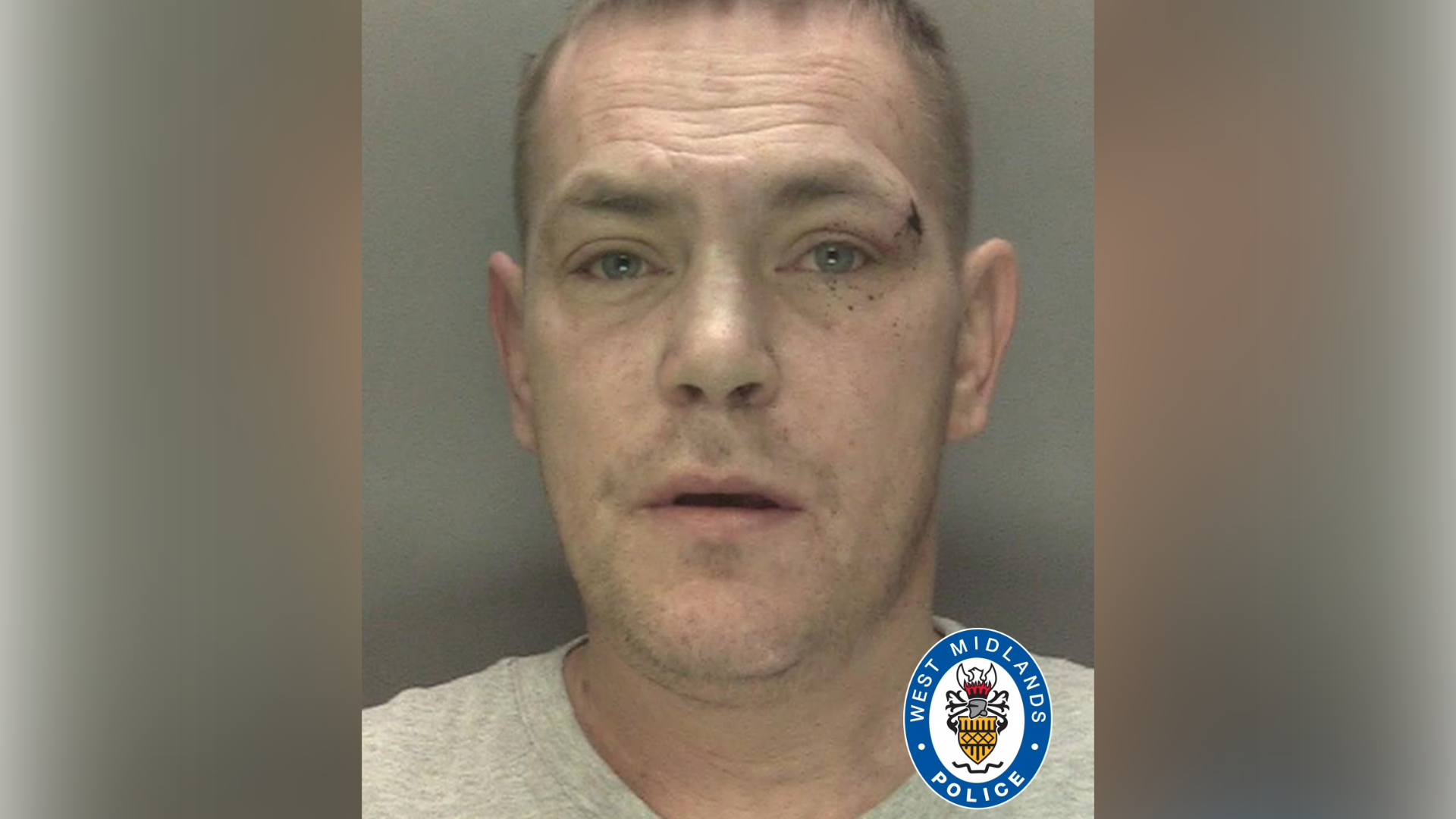 A mug shot of a man who has light brown hair and a grey t-shirt on. He has stubble on his face and a cut next to his left eyebrow