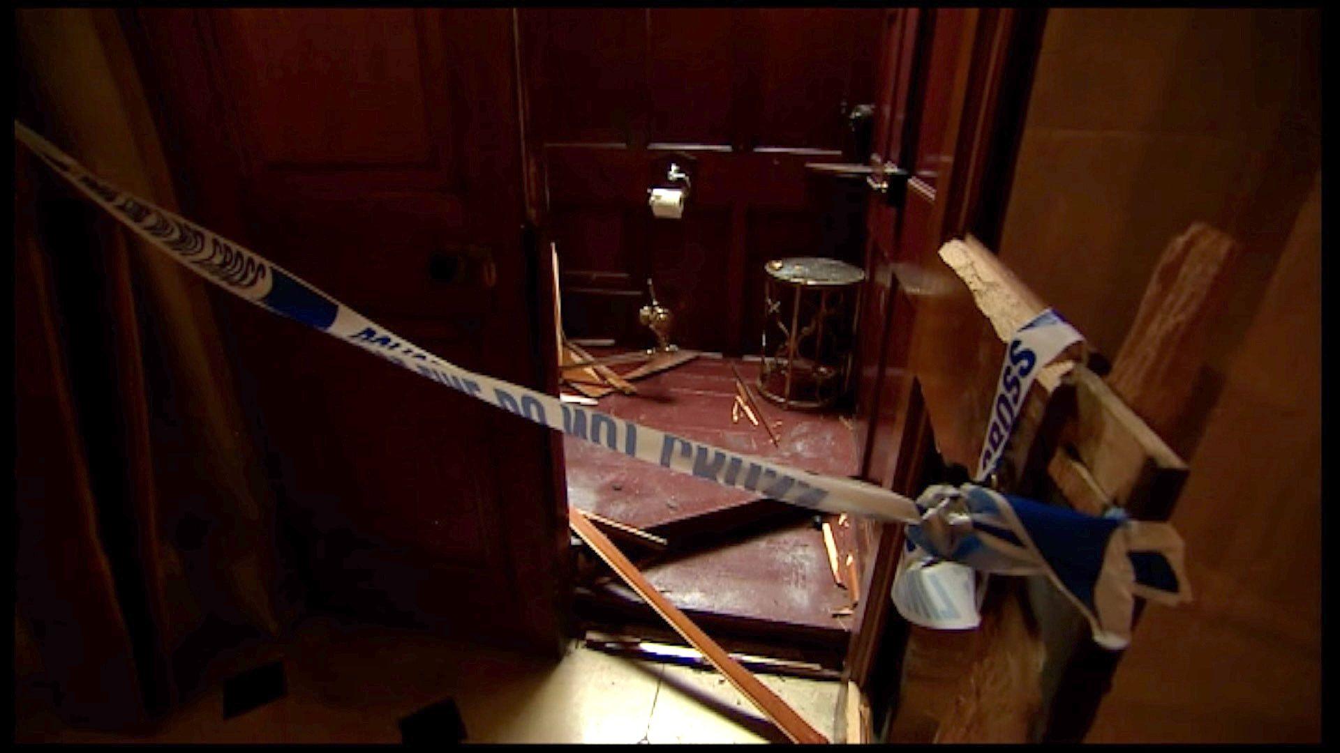 A picture of a small wood pannelled room, with the door open. Police tape is strung across the entrance of the room, and the door is open. Inside there is smashed wood across the floor and some gold pipes lying on the floor. It is a crime scene. 