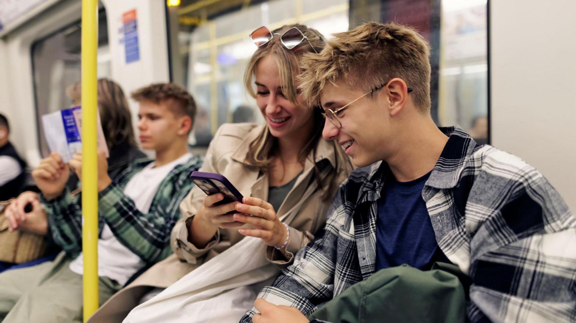A young adult male and a young adult female sit next to each other on a Tube looking at a mobile phone and smiling