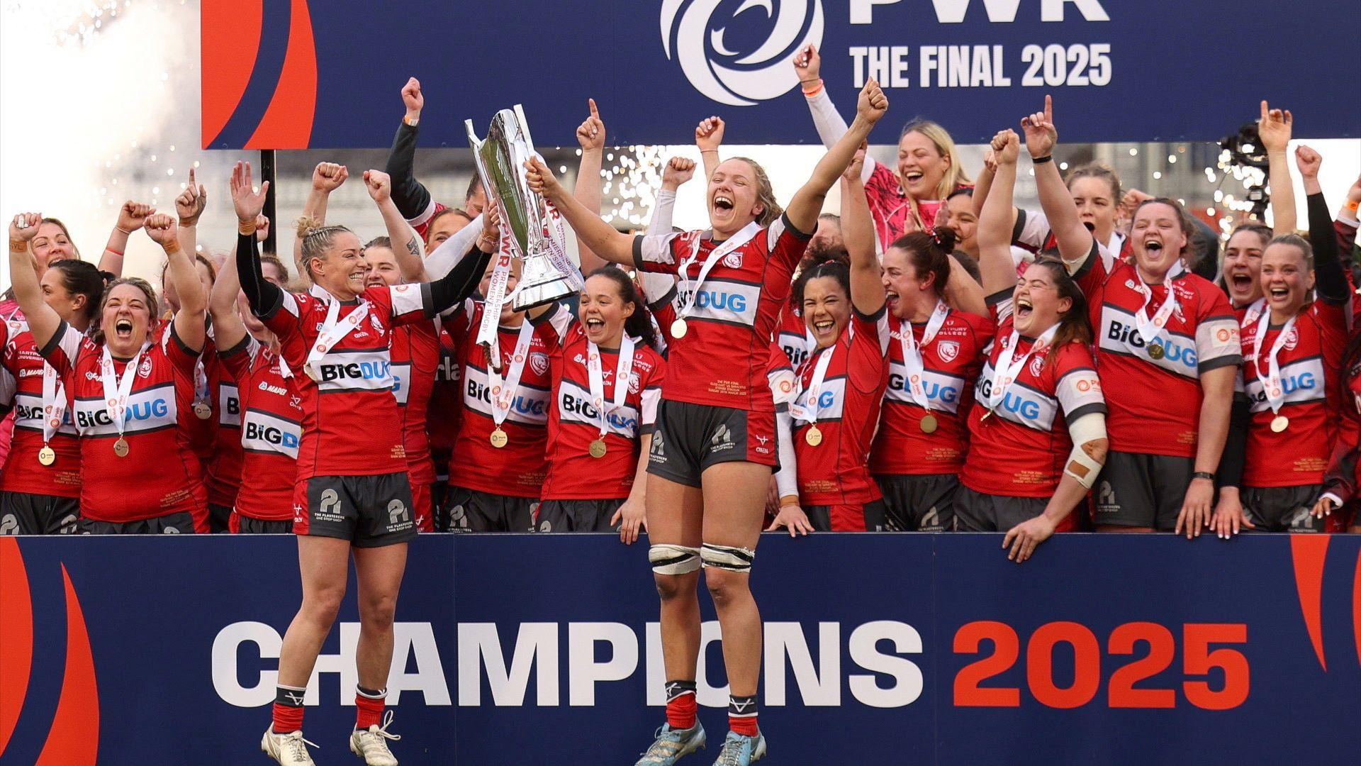 A group of women rugby players celebrating and jumping in the air holding a trophy.